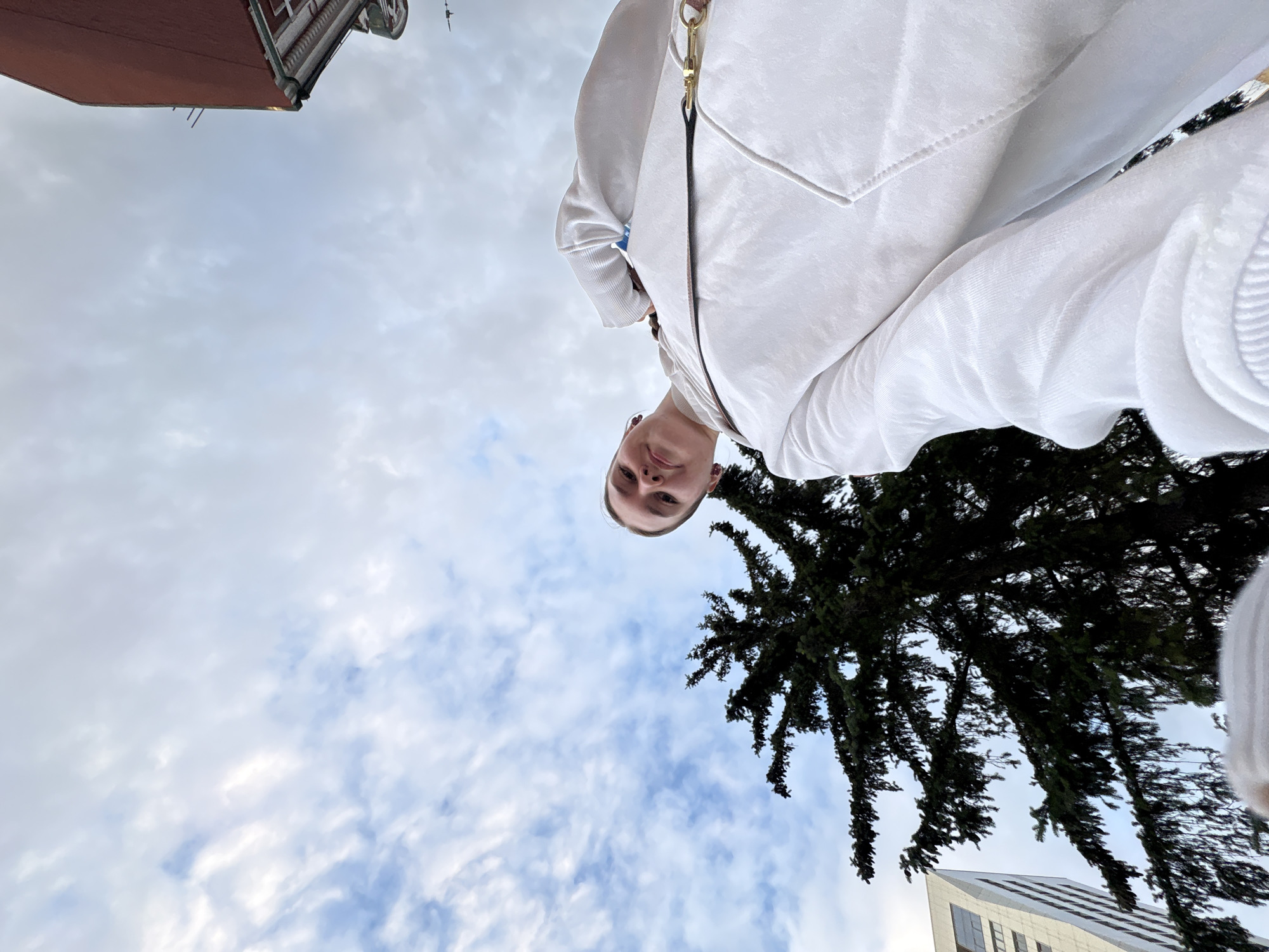 The image is a low-angle selfie featuring a young woman outdoors in the city of Perm, Russia. The woman, positioned in the lower left and center, looks down towards the camera with a subtle smile. She has light skin and wears small red earrings. Her attire consists of a white hooded sweatshirt with a prominent front pocket. A brown strap with black trim and a gold-colored clasp, possibly from a bag, crosses her chest from left to right.
The background primarily displays a cloudy sky, characterized by white and light grey clouds against a pale blue expanse, indicating an overcast day. A large, dark green conifer tree occupies the middle and left portions of the background, with its branches extending widely. To the far left, a portion of a modern white building with visible windows is present. On the far right, a section of a reddish-brown building with a detailed green and white eave is visible, along with what appears to be a small, dark flying object or distant structure against the sky in that area. The perspective of the photograph is from below, looking upwards at the subject and the surrounding urban environment.
