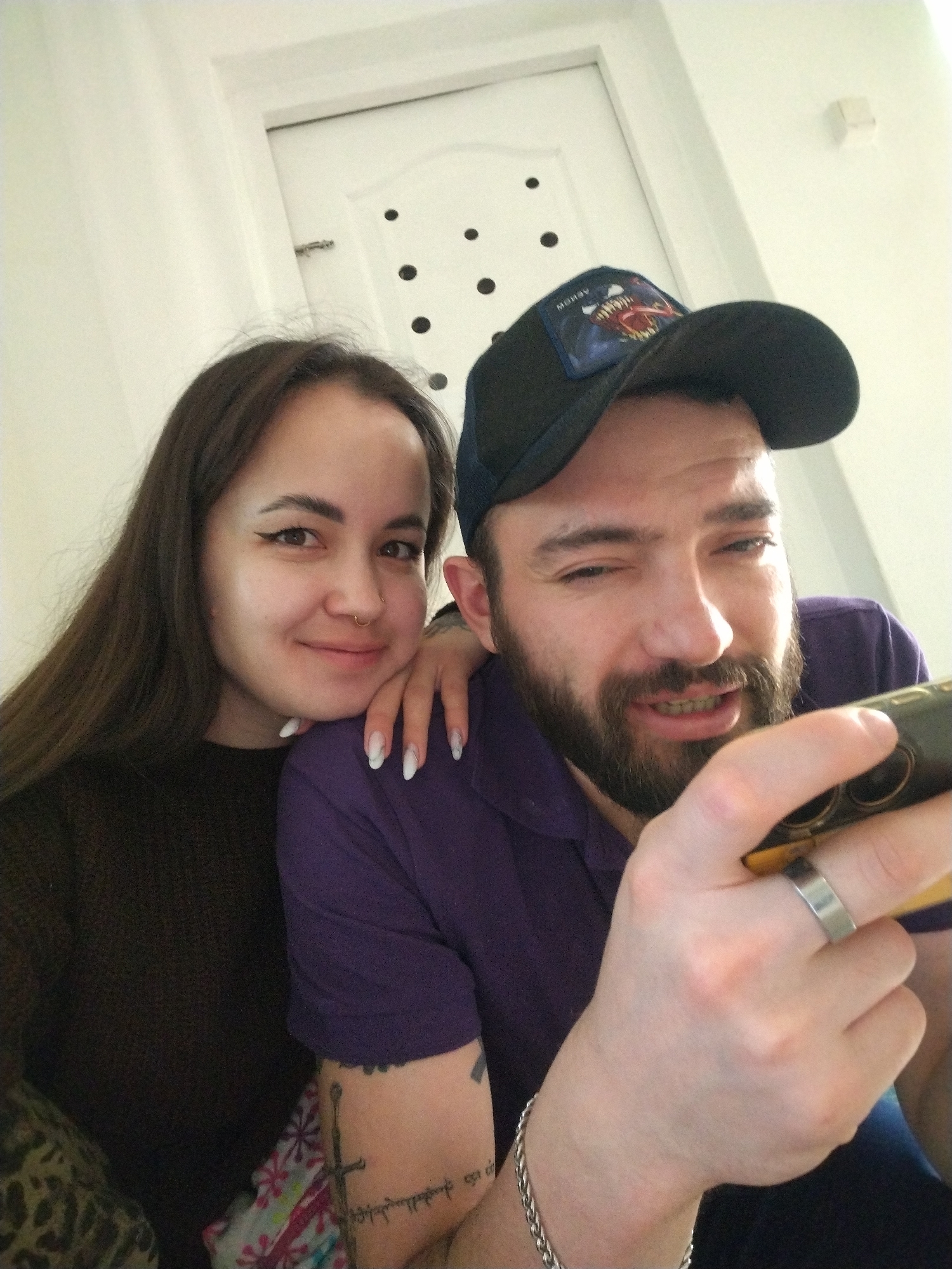 A photograph shows a woman and a man posing together indoors, likely in a domestic setting. The woman, with long dark hair and a nose ring, is on the left, smiling. Her hand rests on the man's shoulder. The man, wearing a dark baseball cap and a purple shirt, is holding a mobile phone and appears to be looking at it intently. He has a beard and a wedding ring on his finger. His left arm is visible, displaying a tattoo of a sword and script. A silver chain bracelet is visible on his wrist. Behind them, a white door with decorative holes and a white wall are visible. There is no visual information in the image to indicate the location as Chaikovsky, Russia.