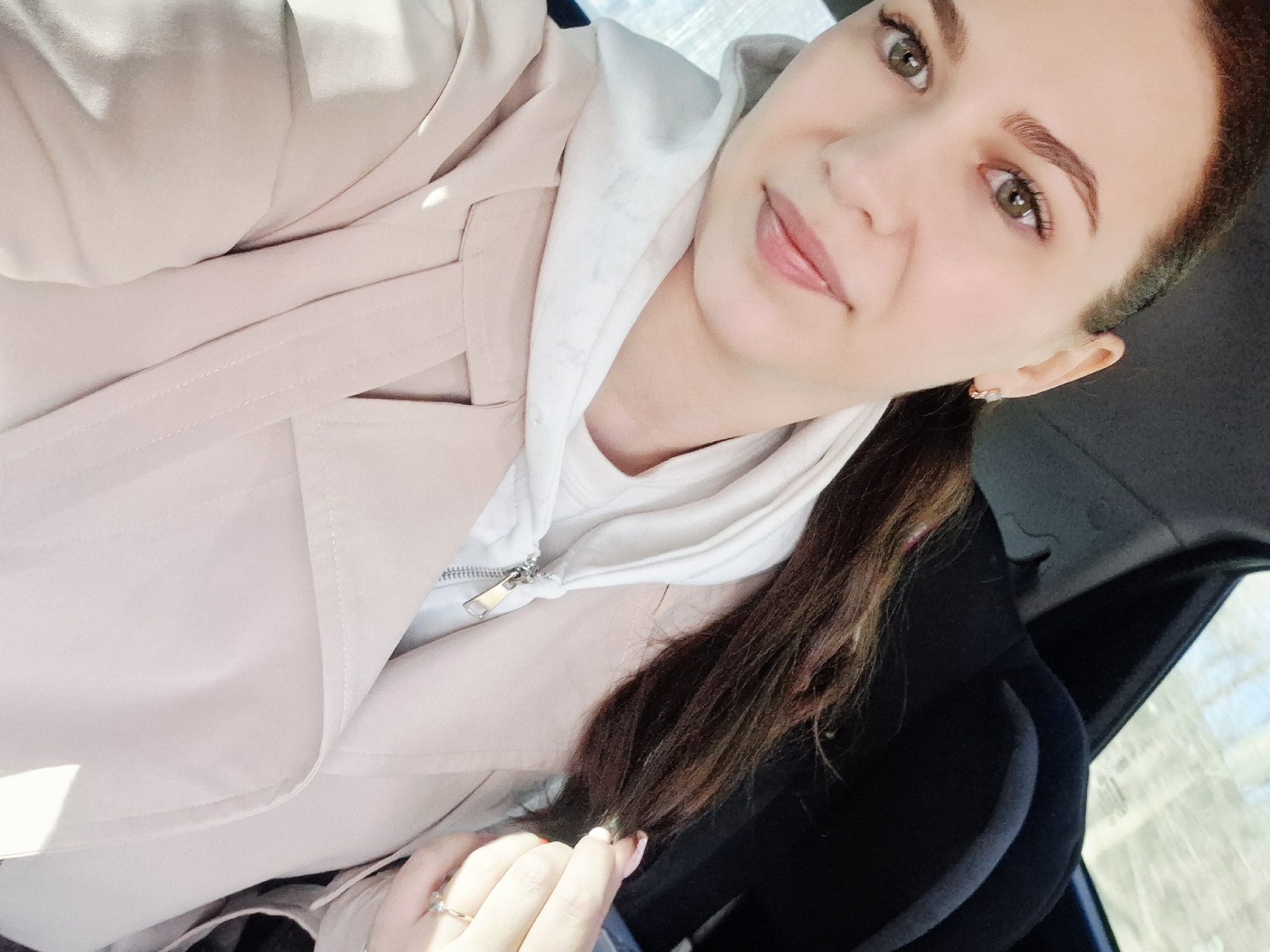 The image is a selfie of a woman. She is wearing a light beige jacket over a white hoodie. Her hair is pulled back into a ponytail. She has light skin, brown eyes, and a ring on her left ring finger. She appears to be sitting in a car, with a black headrest visible in the background. The location is not discernible from the image. It is not possible to confirm if the location is in Chaikovsky, Russia.