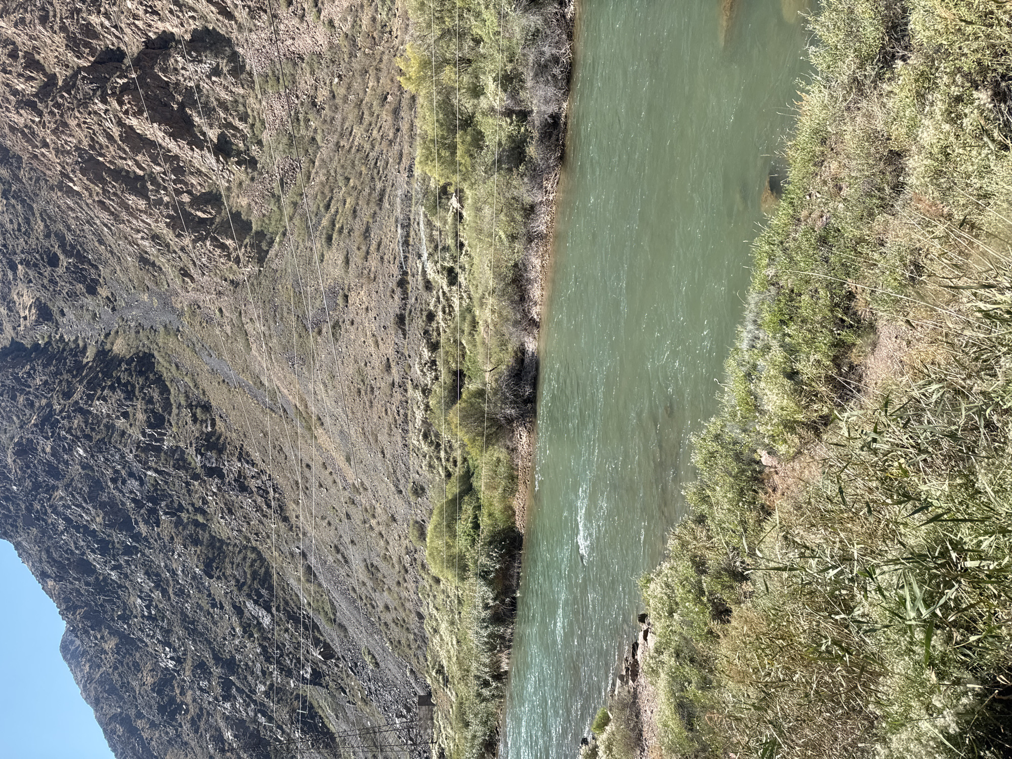 The image shows a wide, turquoise river flowing through a rocky, mountainous landscape. The riverbanks are lined with green vegetation, including tall grasses and shrubs. In the background, a steep, rocky mountain dominates the scene, with sparse vegetation growing on its slopes. The sky is a clear blue. There are power lines strung across the mountain in the background. The location appears to be a natural, undeveloped area.
