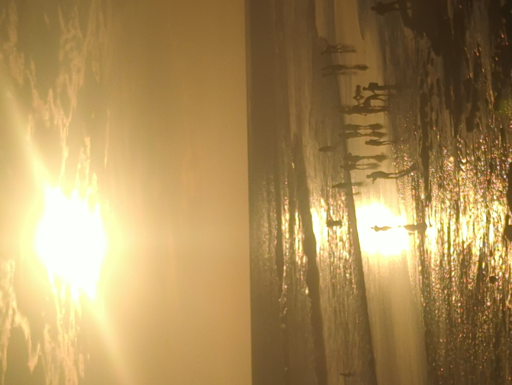 The image captures a sunset scene at North Kuta, Indonesia, featuring a beach filled with silhouetted figures. The dominant element is the bright, obscured sun, emanating intense light. Many people are present, casting dark silhouettes against the glowing light, mostly on the beach. The wet beach reflects light, adding a golden sheen. The background is out of focus, suggesting the sun is the focal point. The overall image has a warm, golden color palette. The photo may have been taken indoors, behind some curtains, or other decor.