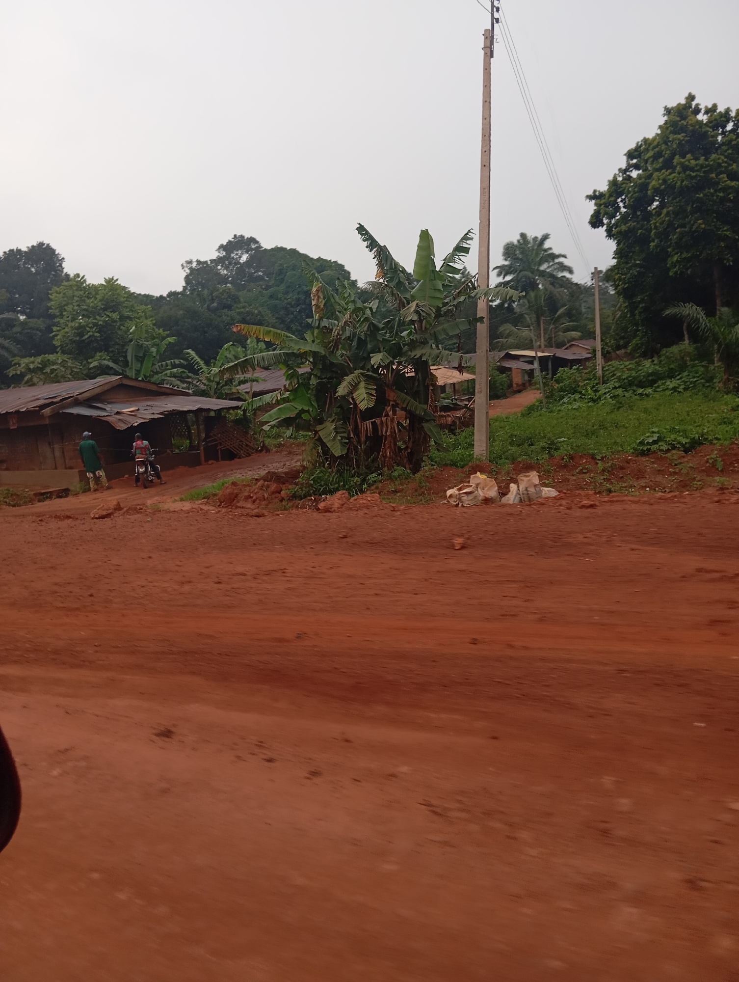 A rural scene in Nsukka, Nigeria, features a dirt road in the foreground, leading towards a cluster of simple buildings and lush vegetation. The sky is overcast. Two individuals are present; one, wearing a green shirt, stands near the buildings, while another is seated on a motorcycle. Several sacks are piled near a utility pole with wires. Banana trees and other foliage are visible, contributing to the tropical environment. The buildings appear to be constructed with wood and corrugated metal roofs. The overall impression is of a modest, somewhat undeveloped settlement within a verdant landscape.