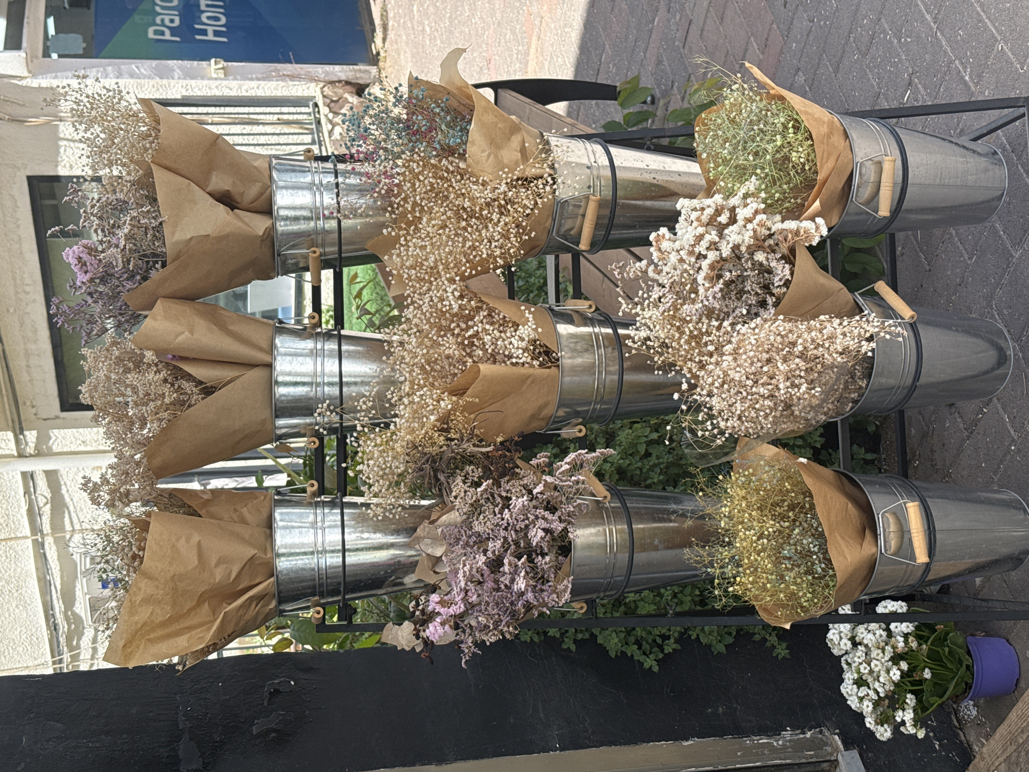 A medium shot, taken outdoors, shows a metal flower display in Raanana, Israel. The display features three rows of galvanized metal buckets, each holding bunches of dried flowers wrapped in brown paper. The top row contains flowers in shades of white, yellow, blue, and brown. The middle row has bouquets of brown and cream-colored flowers. The bottom row displays a mix of purple, yellow, and white flowers. A pot of white flowers sits near the base of the display. The background includes a building with windows and a blue sign with white text. The ground is paved with red bricks. Green plants surround the flower display.