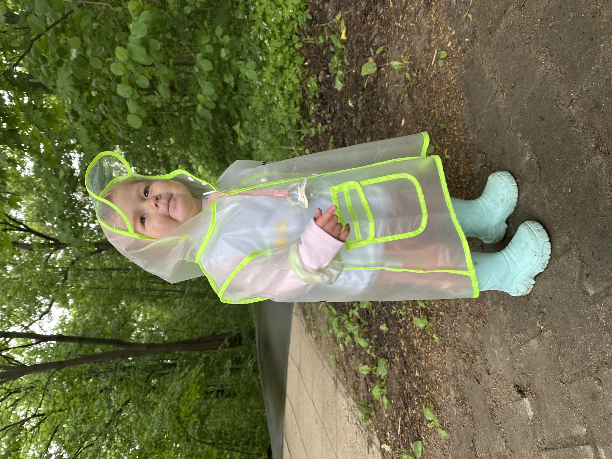 A young child wearing a transparent yellow-trimmed raincoat and blue rain boots stands on a paved surface next to a grassy area. The location appears to be outdoors with green foliage visible in the background. The child is also holding a pacifier. Nizhny Novgorod, Russia cannot be confirmed from the image.