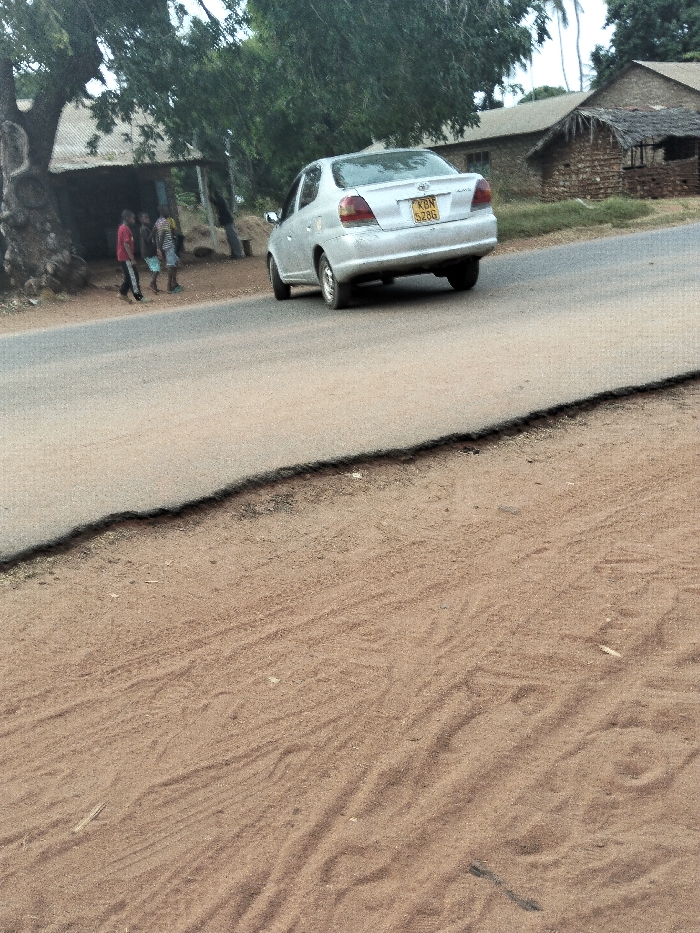 The image captures a roadside scene in what appears to be a rural or semi-rural setting during the daytime. The foreground is dominated by a sandy, light-brown dirt patch with subtle wavy patterns and faint tracks, possibly from vehicles or foot traffic. Above this, an asphalt road runs diagonally across the frame from left to right, showing a distinct, somewhat eroded edge where it meets the dirt.
A silver four-door sedan, resembling a Toyota Vitz or Yaris sedan, is positioned on the asphalt road, moving away from the viewer towards the right side of the frame. Its rear is clearly visible, featuring a yellow license plate that reads "KBN S286".
In the mid-ground, to the left of the road and under a large, leafy tree with a thick trunk, several individuals, likely children or young adults, are standing on the dirt shoulder. They are dressed in casual clothes, with one person in a red shirt and others in striped or light-colored attire. Behind them, partially obscured by the tree, is a simple structure with a corrugated metal roof.
Further in the background, towards the right side of the road, more buildings are visible. One appears to be a larger, stone or concrete structure with a dark roof, while another to its right is a smaller, rustic hut-like building with a thatched or natural material roof. Scattered trees and general vegetation are present throughout the background, indicating a natural environment. The lighting is soft and diffused, suggesting either an overcast day or a time when the sun is not directly overhead, as there are no harsh shadows.
