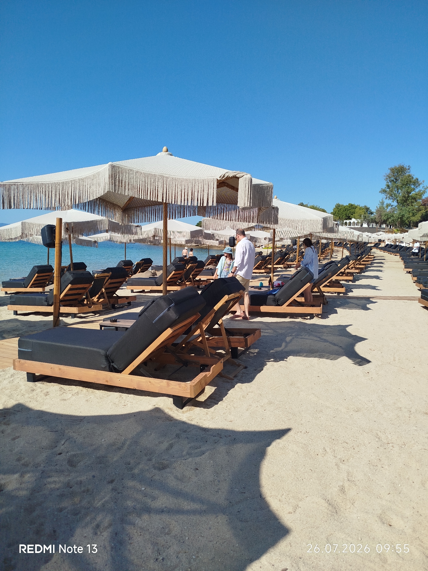 The image depicts a serene beach scene in Sithonia Municipal Unit, Greece, characterized by rows of empty, upscale sun loungers and umbrellas on a sandy shore. The foreground features several black-cushioned sun loungers with wooden frames, some casting distinct shadows on the sand. In the middle ground, a few people are visible, including a man in a white shirt and shorts, and a woman in a blue and white striped shirt, seemingly interacting near the loungers. Further back, more loungers and umbrellas stretch along the beach, leading towards the calm, blue sea. The background reveals lush green trees and some architectural structures, suggesting a resort or a well-developed beach area. The sky is a clear, vibrant blue, indicating a sunny day. A timestamp at the bottom right corner reads "26.07.2026 09:55".