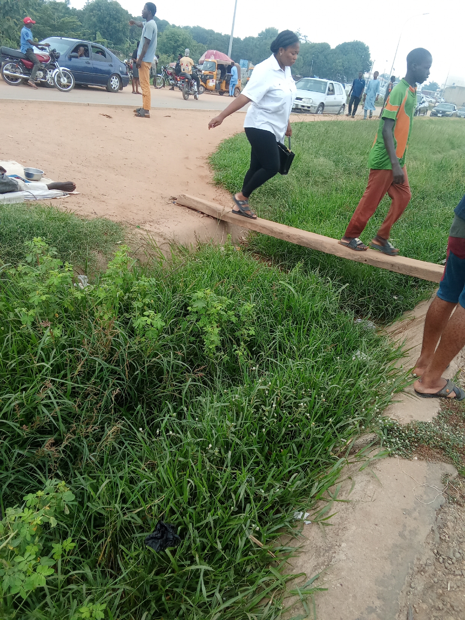 This image depicts a scene in Kagini, Nigeria, likely in a rural or semi-urban area. In the foreground, a wooden plank serves as a makeshift bridge over a ditch filled with lush green grass and some vegetation. Two individuals are walking across this plank. The person on the left, wearing a white shirt and black leggings, is a woman with dark hair pulled back. The person on the right, a younger male, wears a green and orange shirt and brown pants. Their focus is on navigating the unstable crossing.
Further in the background, a dusty road is visible, with several vehicles and people. A blue car is parked on the side of the road, and a person is sitting on a motorcycle in front of it. Another motorcycle, with a rider and passenger, is seen further down the road, alongside a yellow and red structure that appears to be a kiosk or food stall. Several other individuals are walking around this area, and more vehicles are present in the distance. The environment suggests a busy, outdoor setting with elements of both urban and rural life. The overall impression is one of everyday life and movement within the Nigerian town.