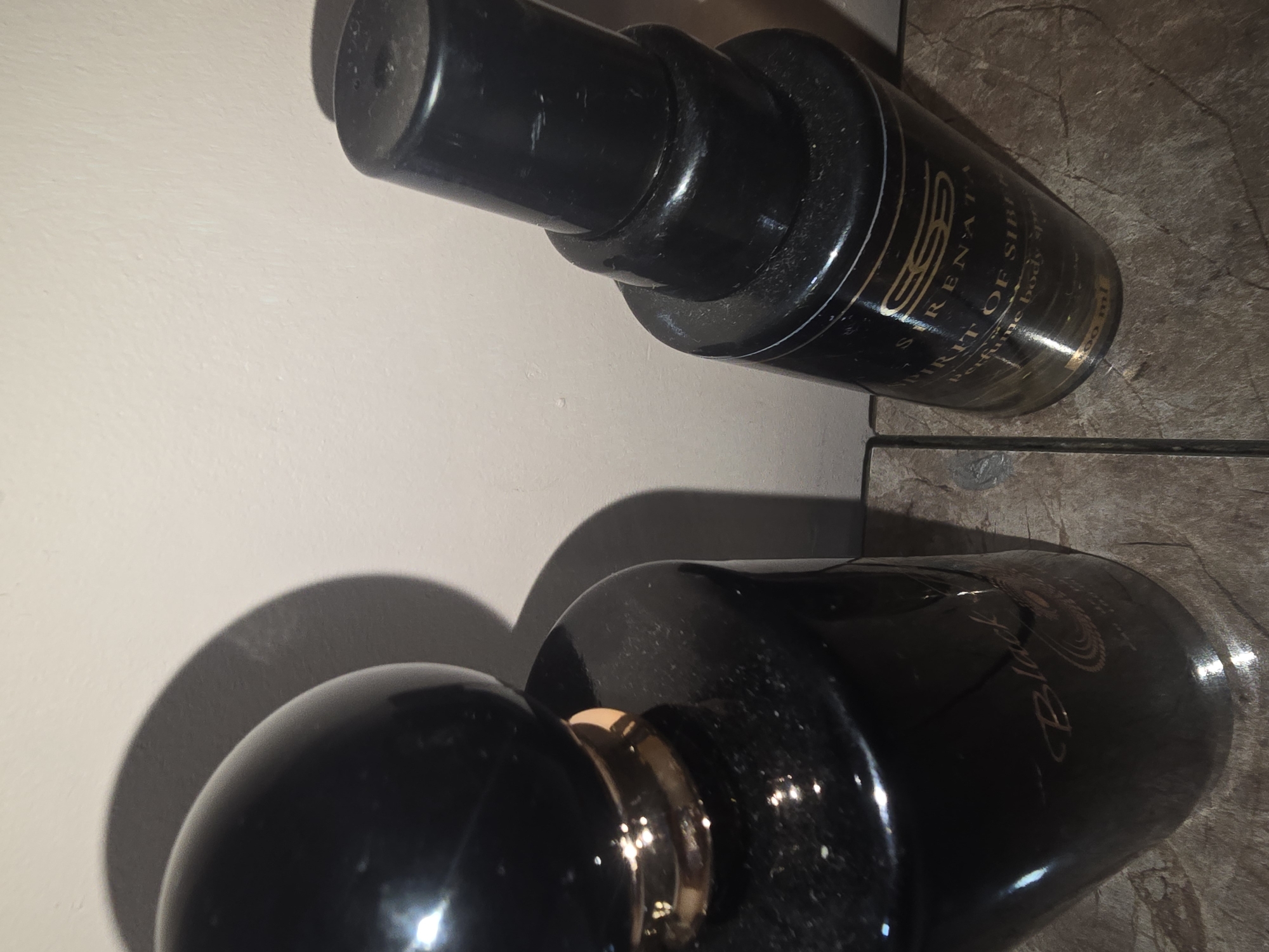 Two black perfume bottles are displayed against a light-colored wall and a tiled surface. The bottle on the left is taller with a rounded, black cap and a gold accent band. The text "Black" is visible in cursive on its dark surface. The bottle on the right is shorter and has a black spray nozzle. Its label is gold and black, with text reading "ESS SIRENATA", "SPIRIT OF SIREN", and "Perfume body spray 100 ml". The lighting creates shadows on the wall. The location of this image is Ulan-Ude, Russia.