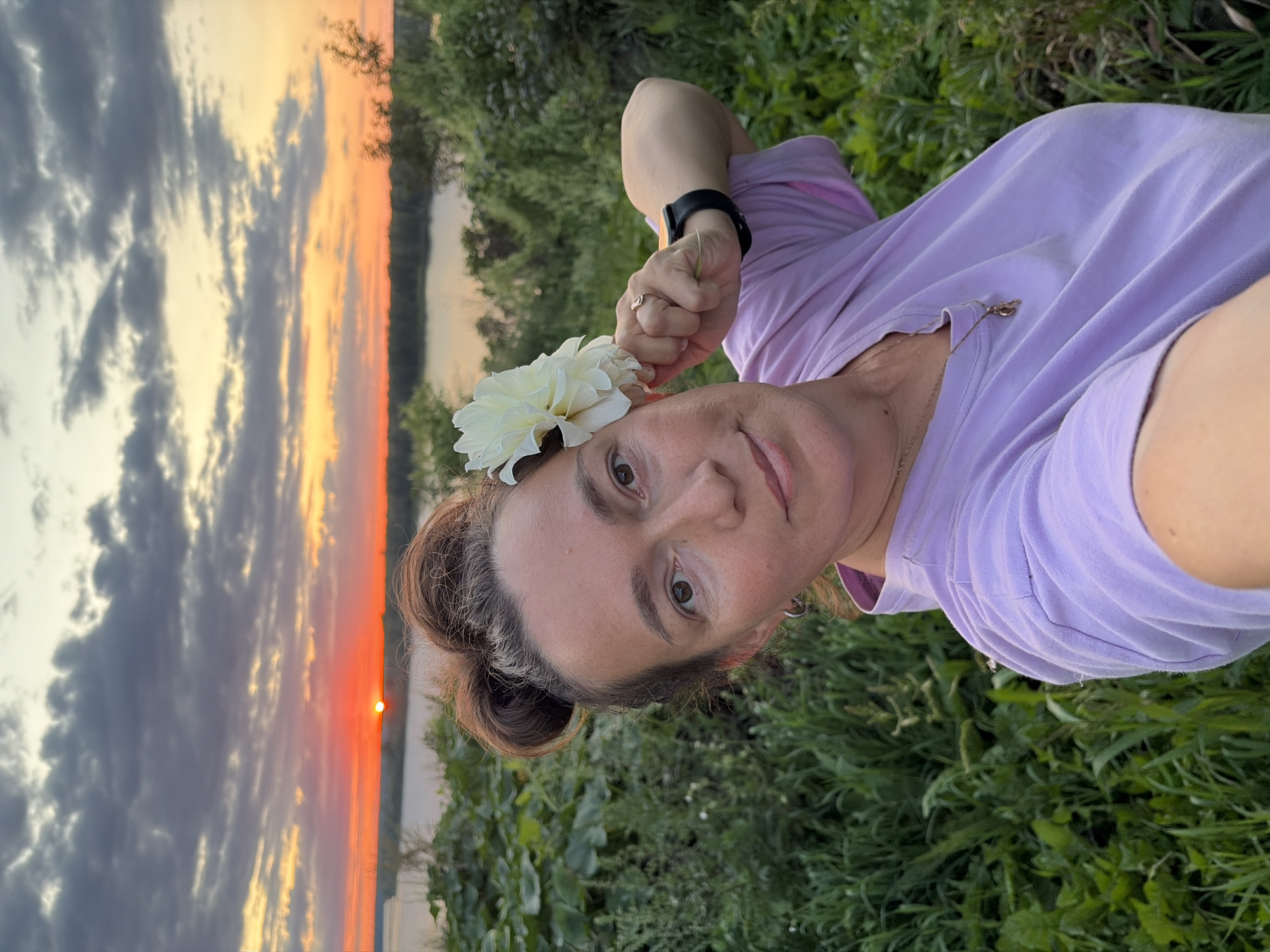 A woman with her hair in two buns is holding a white dahlia to her face, with a scenic sunset and body of water in the background. She is wearing a light purple t-shirt and a gold necklace. She has a ring on her finger and a smartwatch on her wrist. The environment is lush and green, with tall grass and trees surrounding the woman. The sky is filled with dramatic clouds in hues of orange, yellow, and gray. The location appears to be a natural setting, possibly a park or a viewpoint overlooking a river or lake. Olkhovka, Russia.