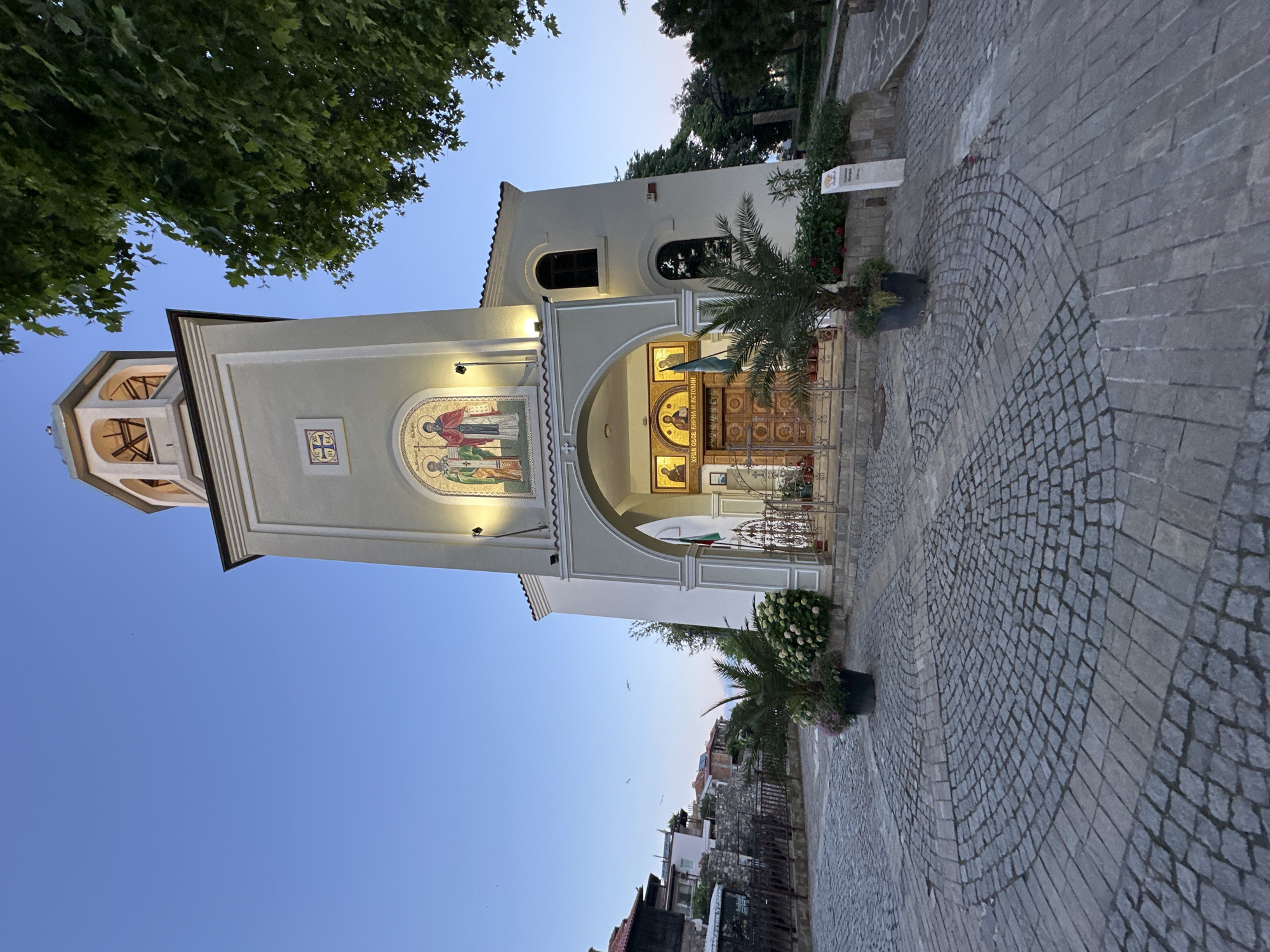 The image depicts the exterior of a church, likely in an Eastern European setting, at dusk. The church features a tall, square bell tower with an arched opening at the top, and is adorned with religious mosaics. The facade of the church has an arched entrance, with ornate wooden doors and decorative metal gates. Potted palm trees and flowering bushes flank the entrance. The ground in front of the church is paved with cobblestones arranged in a circular pattern. In the background, traditional houses with red-tiled roofs are visible, as well as a lush green tree to the right of the frame. The sky is a clear, soft blue, indicating the transition from day to night. There are no people visible in the image.