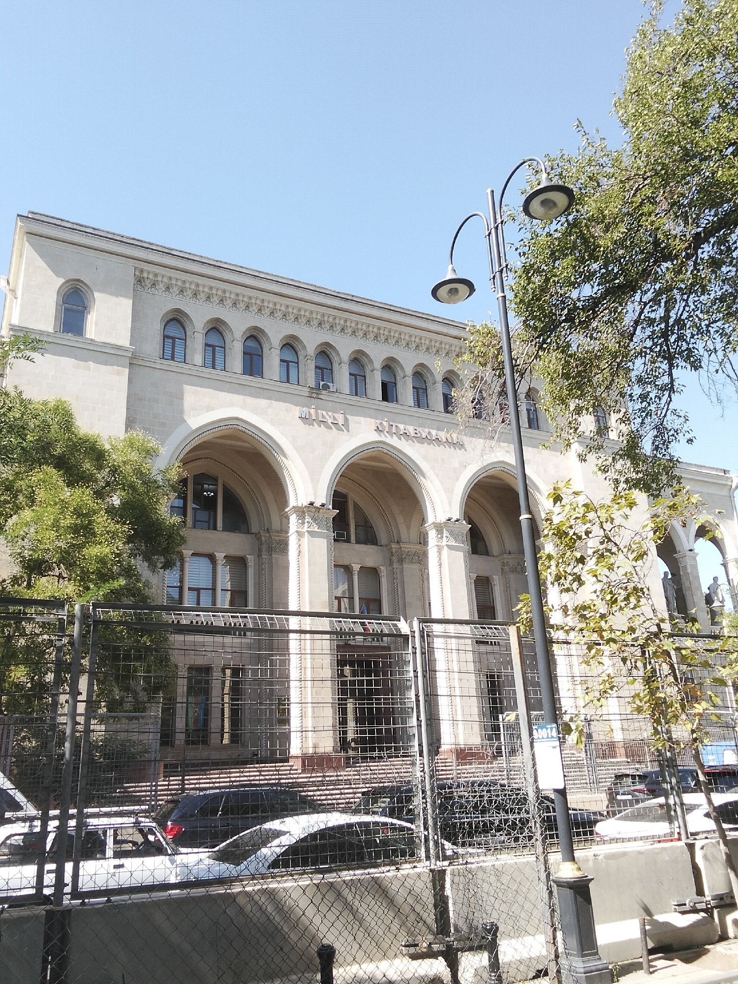 The image captures the exterior of the National Library of Azerbaijan, located in Sabail Raion, Azerbaijan, on a bright, clear day. The facade of the light-colored building is adorned with multiple arched windows and decorative architectural elements. In front of the library, a black metal fence encloses a paved area where several cars are parked, including a white sedan and a darker colored vehicle. Trees with green foliage frame the scene, with one prominently placed near the center of the image. A tall, black lamppost with two lamps stands near the fence, positioned in front of the building. The sky above is a uniform, clear blue.