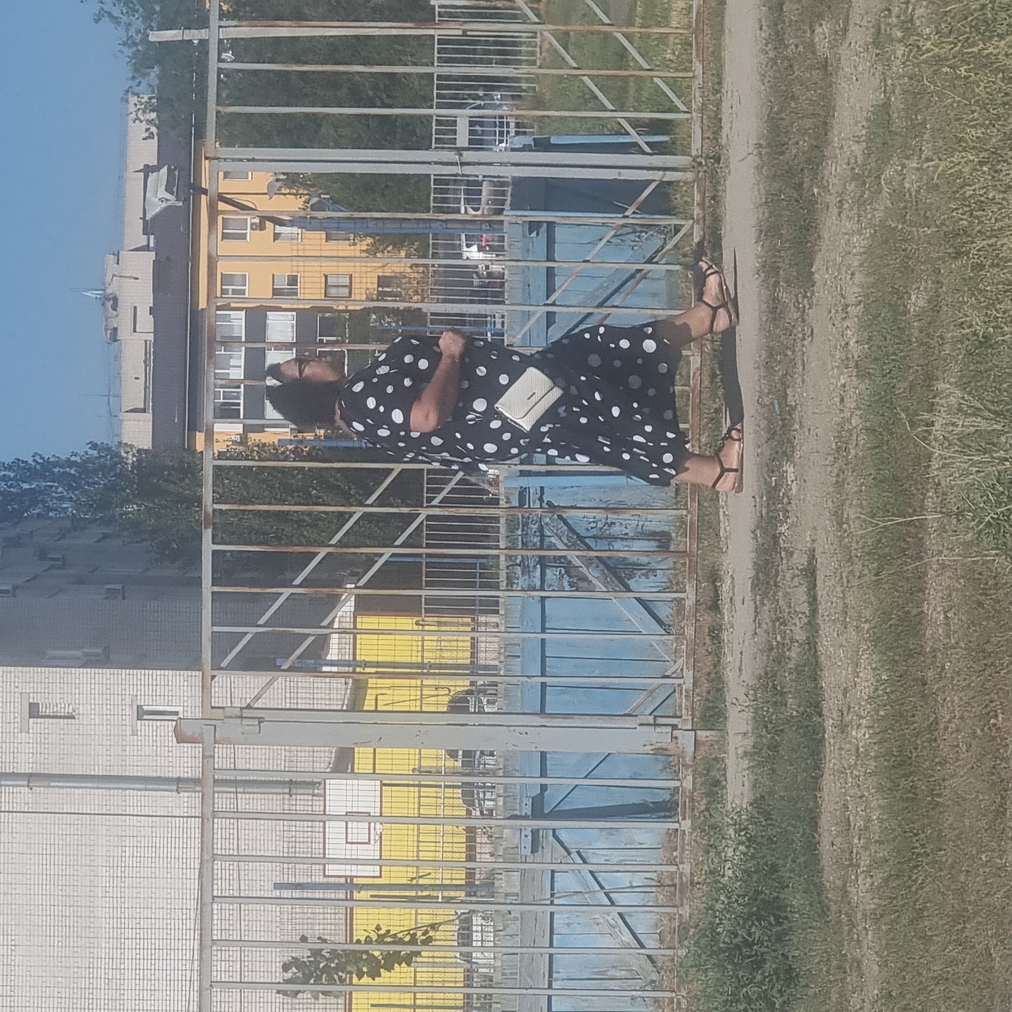 A woman wearing a black and white polka dot dress and black sandals walks past a metal fence. She has dark hair and is carrying a small white purse. In the background, there are buildings, trees, and a blue wooden fence. A basketball hoop is visible on the far left, behind the metal fence. The ground is paved in the foreground and covered with grass and dirt. The scene appears to be an outdoor public space in the city of Aktobe, Kazakhstan.