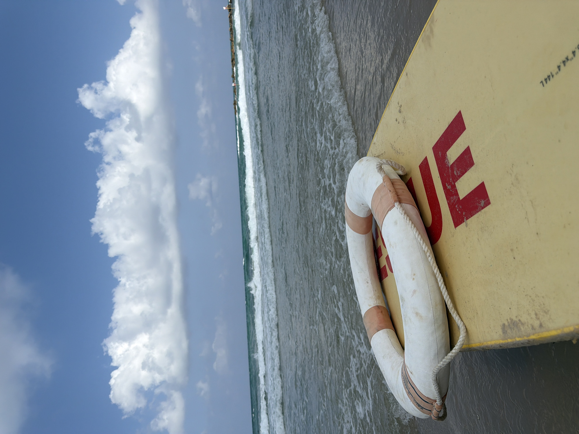 A low-angle, wide shot captures a close-up view of a yellow surfboard with a red "LIFE" inscribed on its surface, resting on the shore of Bat Yam, Israel. A white and orange life preserver is positioned on top of the surfboard. The background showcases a tranquil sea with gentle waves rolling onto the sandy beach. In the distance, a breakwater juts out into the water, with a red and white beacon visible at its end. The sky above is a clear blue, dotted with fluffy white clouds. The scene evokes a sense of calm and preparedness for beach activities.