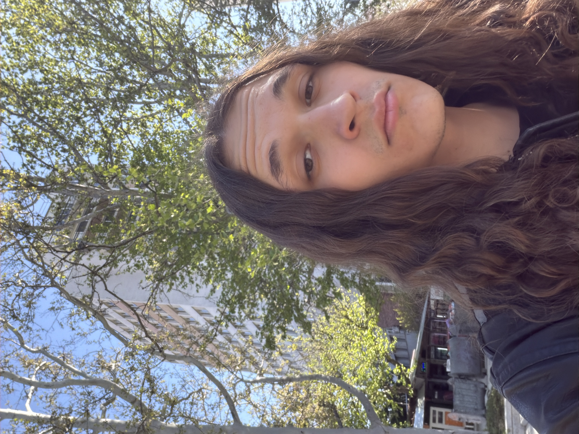 The image is a selfie-style portrait of a young person with long, wavy brown hair, looking directly at the camera. The person has a neutral to slightly contemplative expression, with subtle forehead lines and light stubble on the chin and upper lip. The subject is wearing a dark jacket or shirt, partially visible on the right.
The background depicts an outdoor urban environment in Plovdiv, Bulgaria. Lush, bright green leaves on tree branches fill the upper left and central portions of the frame, indicating springtime or early summer. A clear blue sky is visible between the foliage. Behind the trees, a tall, light-colored building with multiple windows can be seen. Further in the background, to the right, other indistinct buildings or structures contribute to the city atmosphere. The bright, clear sunlight suggests a sunny day, likely during daytime hours. No discernible text is visible within the image.
