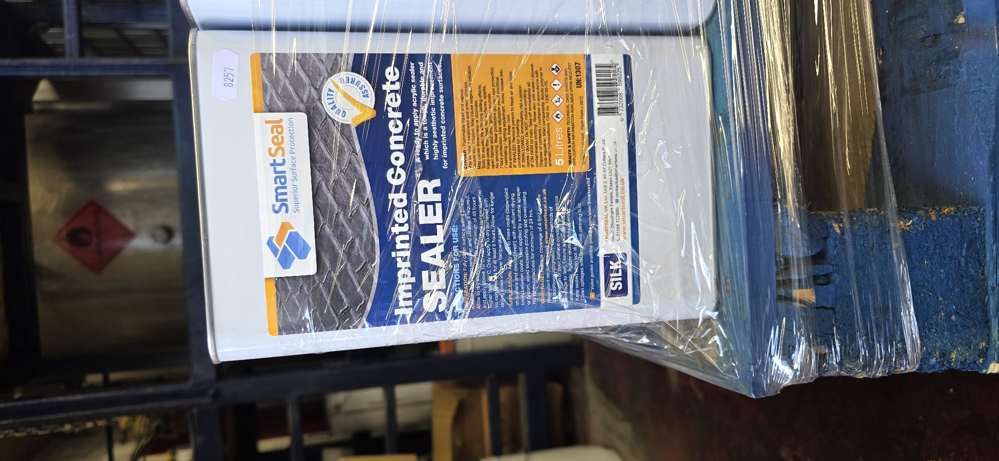 The image shows a white metal container of SmartSeal Imprinted Concrete Sealer. The container is wrapped in plastic and is sitting on a blue wooden pallet. The pallet is partially visible and appears to be in a warehouse or storage facility. The background is out of focus but shows industrial shelving and metal structures. A hazard symbol is visible on a metal structure in the background. The container has a label with product information and instructions for use. The location in Arun, United Kingdom, is not discernible from the image.