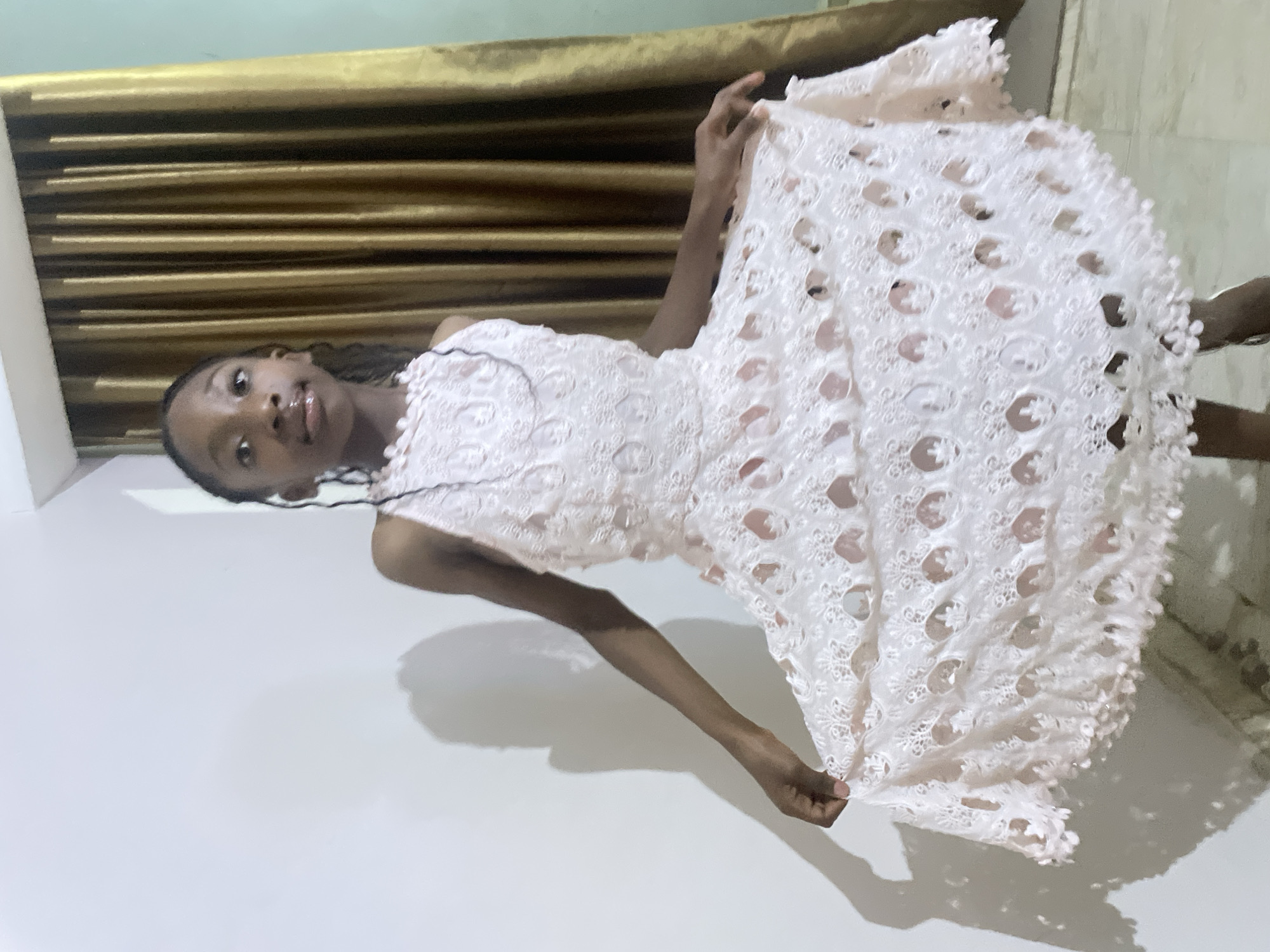 A young person with dark skin, presumably female, is seen indoors. The location is unknown. The person is holding up a light pink or white lace dress. The background features a gold-colored curtain or wall covering. The person is facing the camera with a neutral expression.