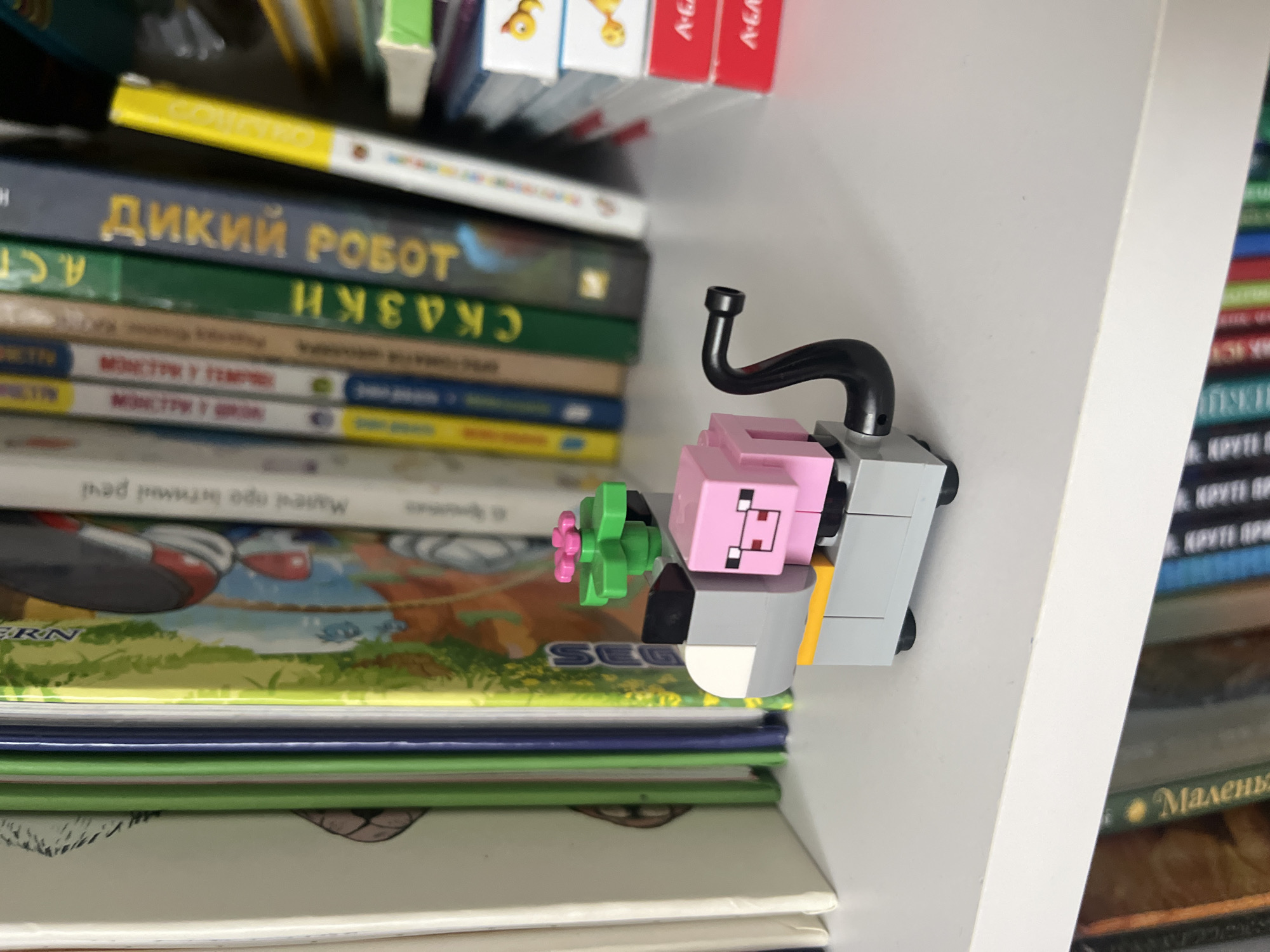 In Onokivtsi, Ukraine, a Lego figurine of a pink pig with a black exhaust pipe is placed on a white shelf. The figurine is positioned in front of a row of colorful books, some of which have visible titles and illustrations. The pig's head is pink, with black eyes and a snout. It sits on a gray base with yellow accents. A green plant with pink petals sits on top of the pig's back. The background consists of a bookshelf filled with various books, and some stack of books on the right side of the image.