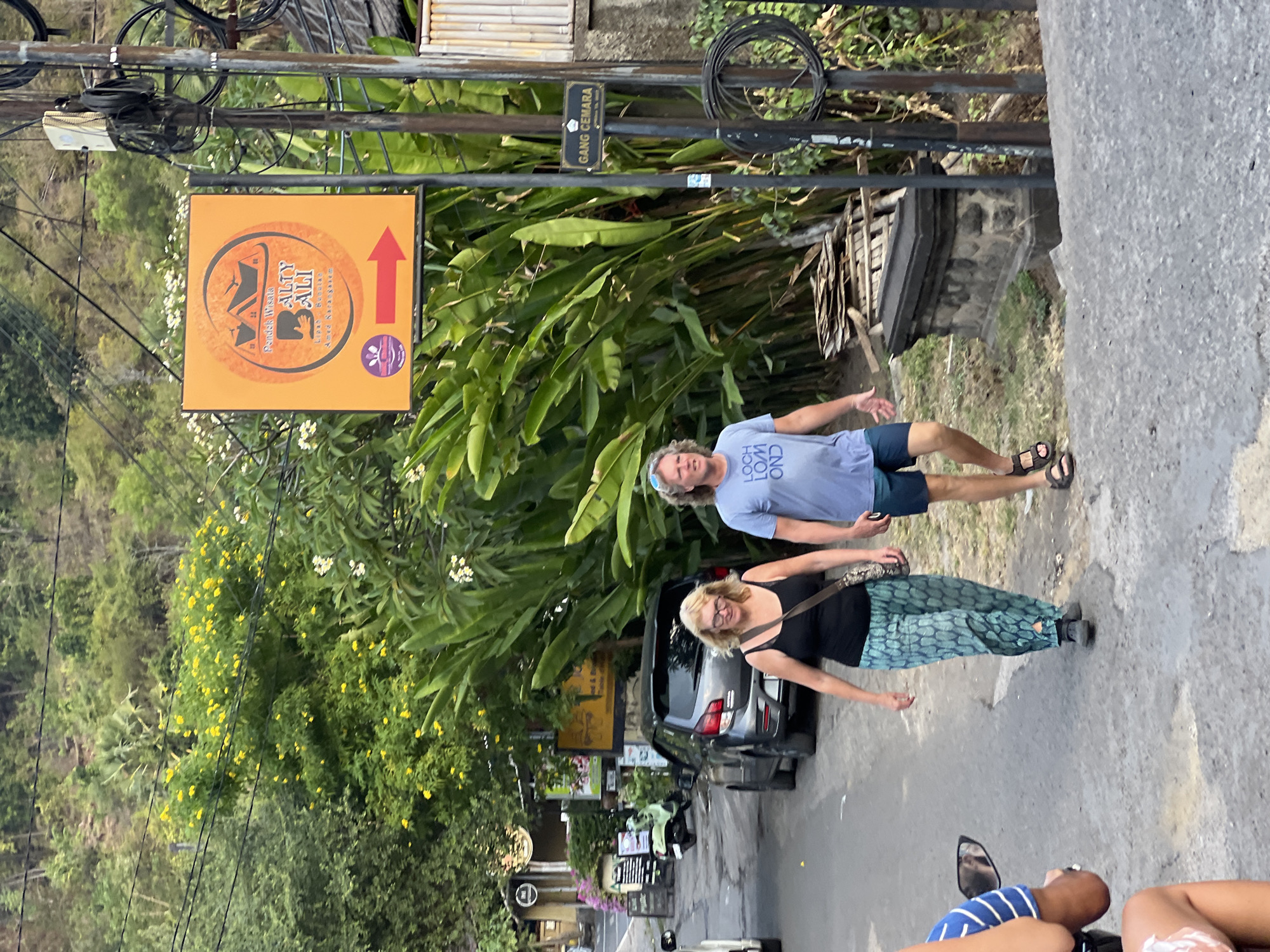 A man and a woman walk down a paved road in Abang, Indonesia. The woman is on the left, wearing a black tank top and green patterned pants. The man is on the right, wearing a blue t-shirt and dark shorts. Behind them, a dark gray car is parked on the side of the road. A sign with orange and yellow branding is mounted on a pole to the right of the road. The sign features a graphic of boats and text, and a red arrow points to the right. A smaller black sign reads "GANG CEMARA". Lush green foliage and trees surround the area.