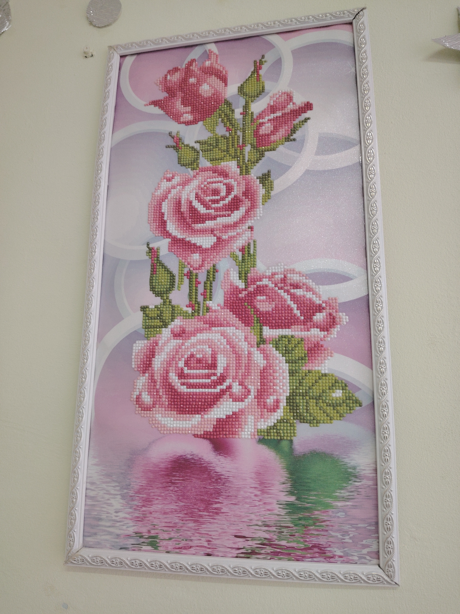 A framed diamond painting featuring a bouquet of pink roses hangs vertically on a plain, light-colored wall. The artwork depicts three prominent, fully bloomed pink roses and several rosebuds, surrounded by green leaves and stems. The roses are rendered in a pixelated style, characteristic of diamond painting, using various shades of pink and white for the petals and different greens for the foliage. The background transitions from a soft pink to a lighter hue, with subtle white, swirling, ribbon-like or circular patterns.
The bottom portion of the image shows the roses and their leaves reflected in a wavy, water-like surface, creating a mirror effect with distorted colors and shapes. The frame itself is white, made of a material with a slightly textured or embossed decorative pattern that resembles connected floral or abstract motifs. In the upper left corner of the image, two small, circular, sparkly decorations are partially visible on the wall above the frame, along with a small white object near the top edge of the frame. The setting appears to be an interior space in Sarapul, Russia, where the artwork is displayed as decorative wall art. No specific activity is taking place, and no text is visible.