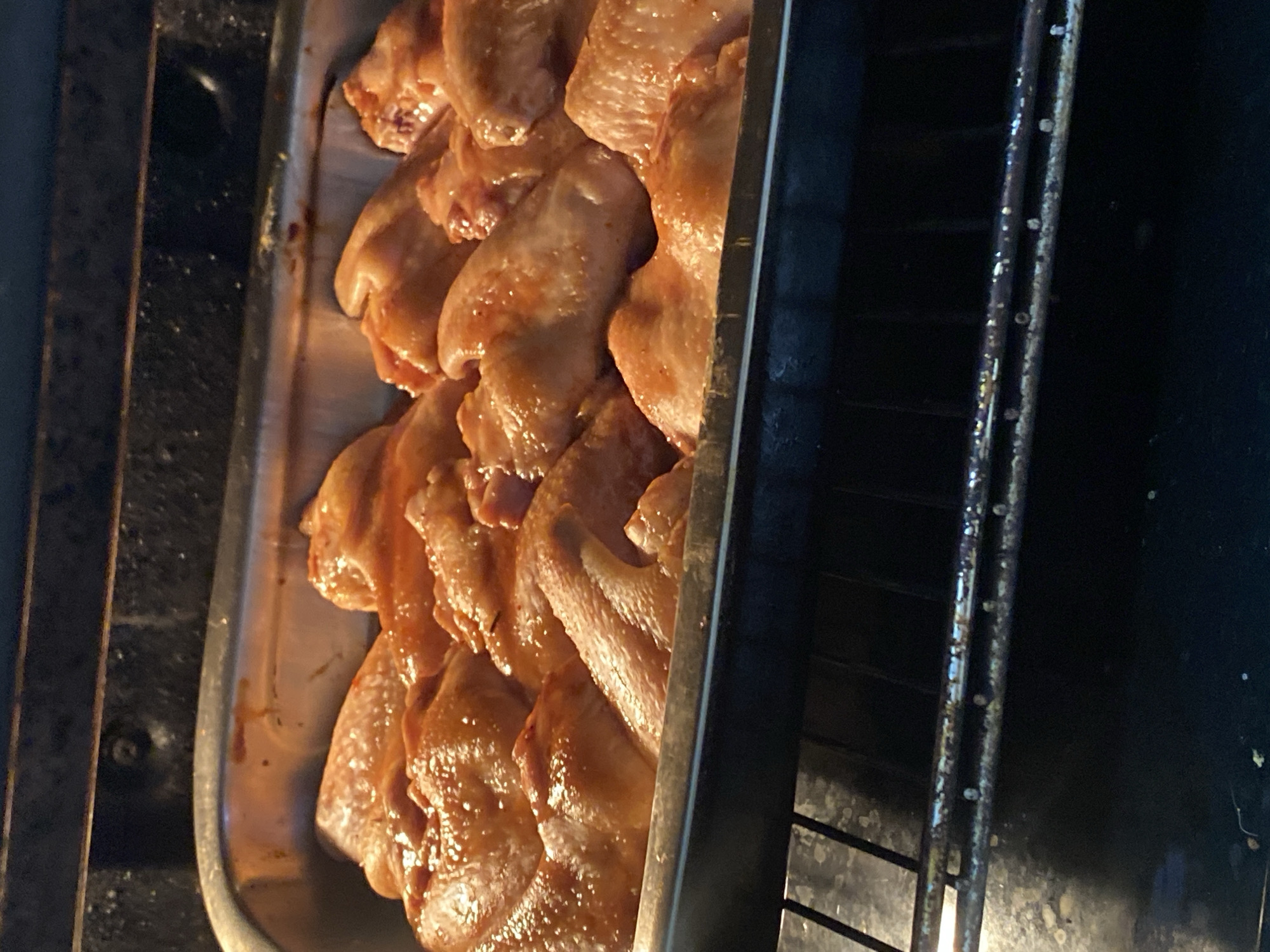 Chicken wings are being roasted in a baking pan inside an oven. The wings appear to be coated in a reddish-orange sauce, likely a marinade or seasoning. The baking pan is silver and is positioned on an oven rack. The interior of the oven is visible, with heating elements and the dark metal walls of the oven. The lighting suggests the flash from a camera or the interior light of the oven. This image was taken in Chornomorsk, Ukraine.