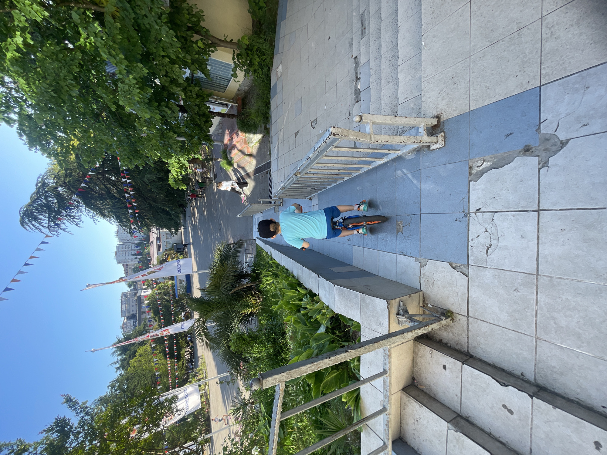 An elevated, high-angle outdoor shot captures a public pedestrian area in Sochi, Russia, under clear blue skies and bright daylight. In the immediate foreground, a young child, viewed from behind, wears a light blue t-shirt, blue shorts, and a black baseball cap while seated on a small orange and blue bicycle. The child is positioned on a tiled landing between two descending flights of concrete stairs. Both the stairs and the landing display significant deterioration, with numerous chipped, cracked, and missing light gray and blue tiles. Weathered and rusted metal railings, originally white, flank the stairs.
Beyond the stairs, a wide paved path or plaza extends into the mid-ground. Along this path, a woman with light hair, dressed in a white top and dark pants, walks towards the right, carrying a dark bag. Further back and to the left, another individual in athletic wear is seen running. The area is adorned with decorative triangular pennants in red, white, and blue, strung across trees and poles. Several large white flags bearing the Cyrillic word "ОБЪЕДИНЕНИЕ" (ASSOCIATION/UNION) in red and black text are visible on poles to the left. Lush green vegetation, including a large palm tree and various other trees, populates garden beds and the surrounding landscape. In the background, multiple multi-story buildings are visible, nestled amidst dense greenery on a distant hillside. A light yellow building with distinctive slanted windows is situated to the right of the paved path at the base of the stairs.
