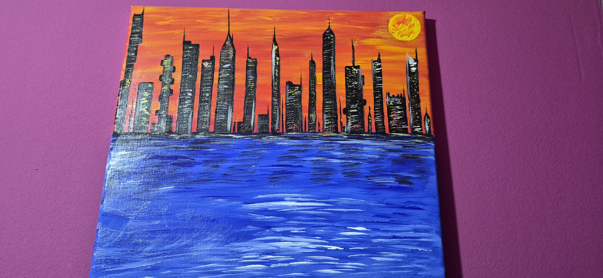 This is a painting of a cityscape with a sunset and a body of water in the foreground. The cityscape consists of numerous skyscrapers of varying heights and designs, painted in black and gray with yellow accents to represent lit windows. The sky is a vibrant gradient of orange and red, transitioning from a bright yellow-orange near the horizon to a deeper red at the top. A circular yellow sun is visible in the upper right corner of the sky. The foreground depicts a body of water, painted in various shades of blue with white highlights to simulate waves and reflections. The painting is displayed against a textured purple wall. There is no indication of the city of Lom, Bulgaria, in this image; it is a generalized representation of a city.