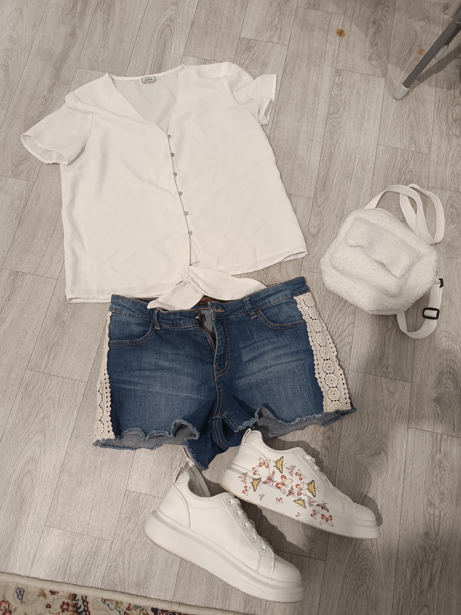 A flat lay image shows a white short-sleeved blouse with a v-neck and button closure, tied at the waist. Below the blouse are denim shorts with lace detailing on the sides and a frayed hem. A small, fluffy white crossbody bag with a flap closure and strap is positioned to the right of the shorts and blouse. Completing the outfit are a pair of white sneakers with a platform sole, decorated with a colorful butterfly print. The items are arranged on a light-colored wooden floor, which shows some minor scuff marks. The scene appears to be an indoor setting, possibly a domestic space. There are no people or actions depicted in the image. The image is taken from a top-down perspective. The location within the city administration of Taraz, Kazakhstan is not discernible from the visual information provided in the image.