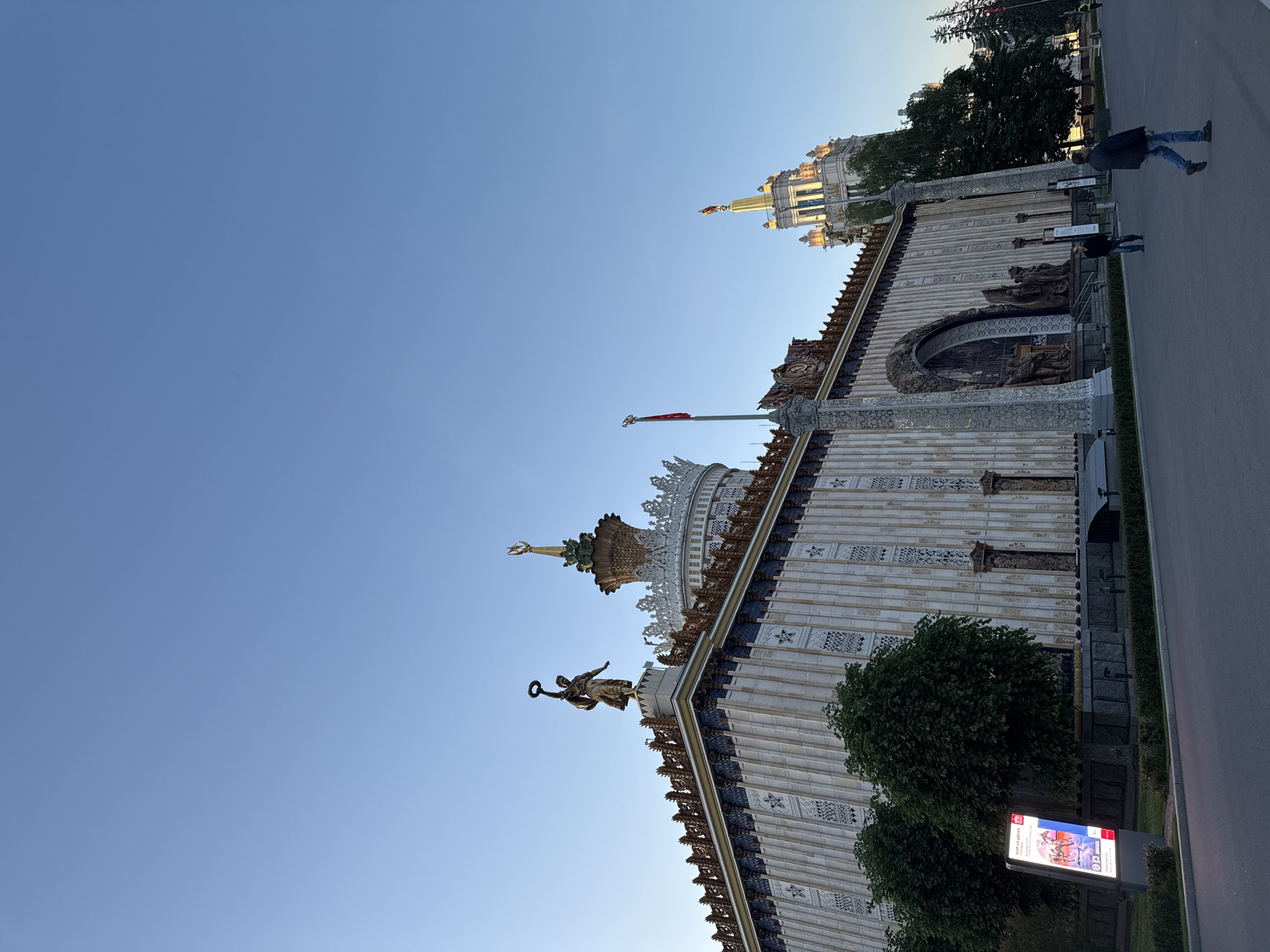 The image displays a grand and ornately decorated building under a clear blue, late afternoon sky. The architectural style, characterized by its monumental scale, intricate detailing, multiple domes, and golden accents, strongly suggests a location like the Exhibition of Achievements of National Economy (VDNKh) in Moscow, Russia.
The main subject is a large, light-colored building with a textured facade and numerous decorative elements, including sculpted friezes and patterned panels. Several golden statues adorn the building's roofline and domes. One prominent golden figure holding a wreath is visible, and another golden statue crowns a central spire. A red flag flies from a flagpole positioned on the building. In the background, another distinct building with domes and spires is partially visible, indicating a complex of similar structures.
To the right of the building, a paved pathway runs alongside a grassy area with green trees. Two individuals are walking on this pathway; one person wears a dark jacket and blue jeans, while a smaller figure follows behind. The natural lighting suggests the golden hour, with the sun casting a warm glow on the building and statues.
Near the pathway and partially obscured by foliage, a modern, illuminated digital sign or billboard displays colorful images, likely advertising an event or performance, with some unreadable Cyrillic text. The scene is calm, depicting a public space with visitors enjoying the grand surroundings during favorable weather.
