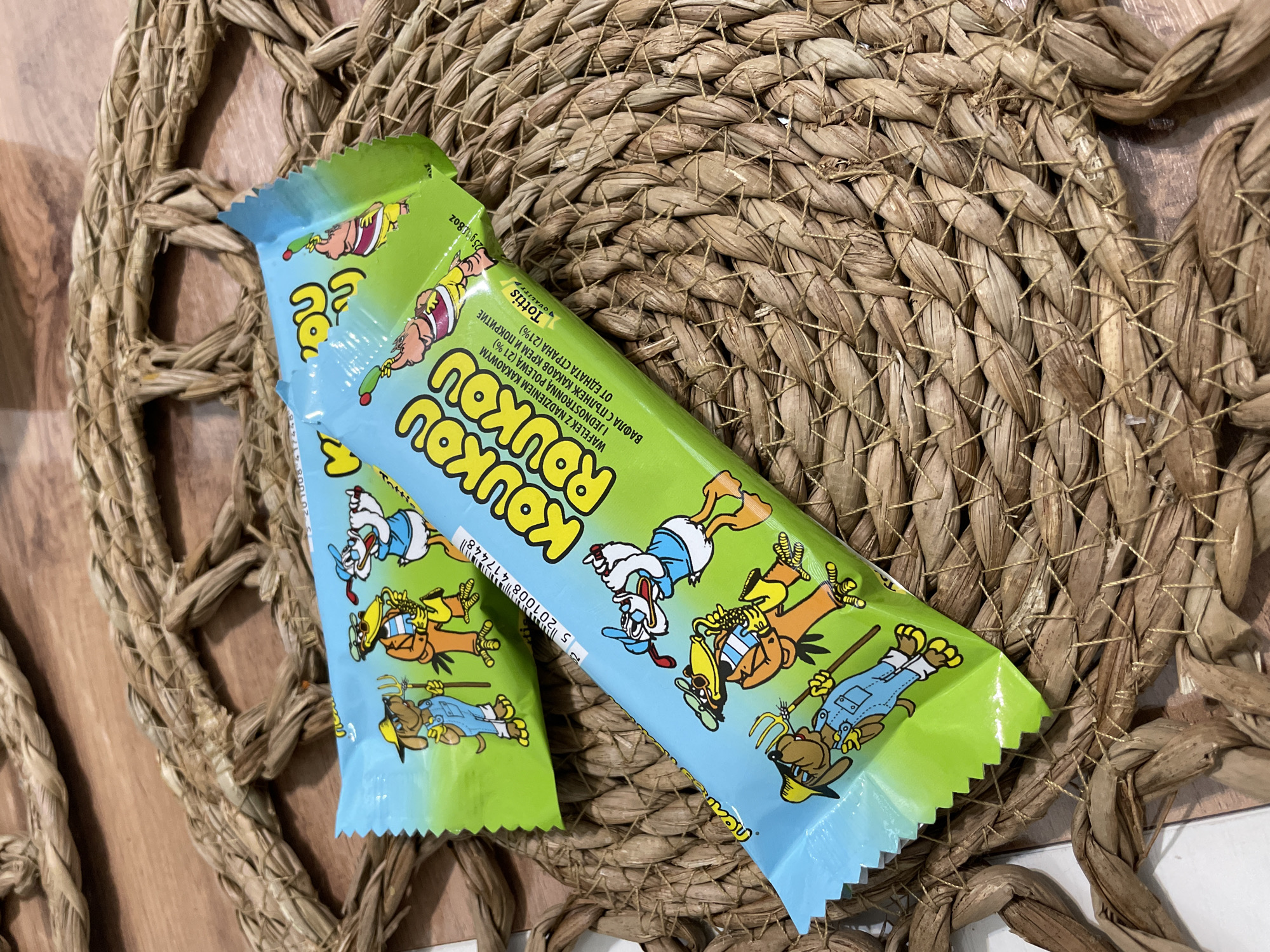 Two "Roukou Roukou" wafer bars with cartoon character illustrations are positioned on a circular woven mat made of natural fibers. The mat is placed on a surface with a mixed texture: part wood and part white. The wrapper's text is not in English, but the text does say "Tottis" which is a product of Chaikovsky, Russia.
