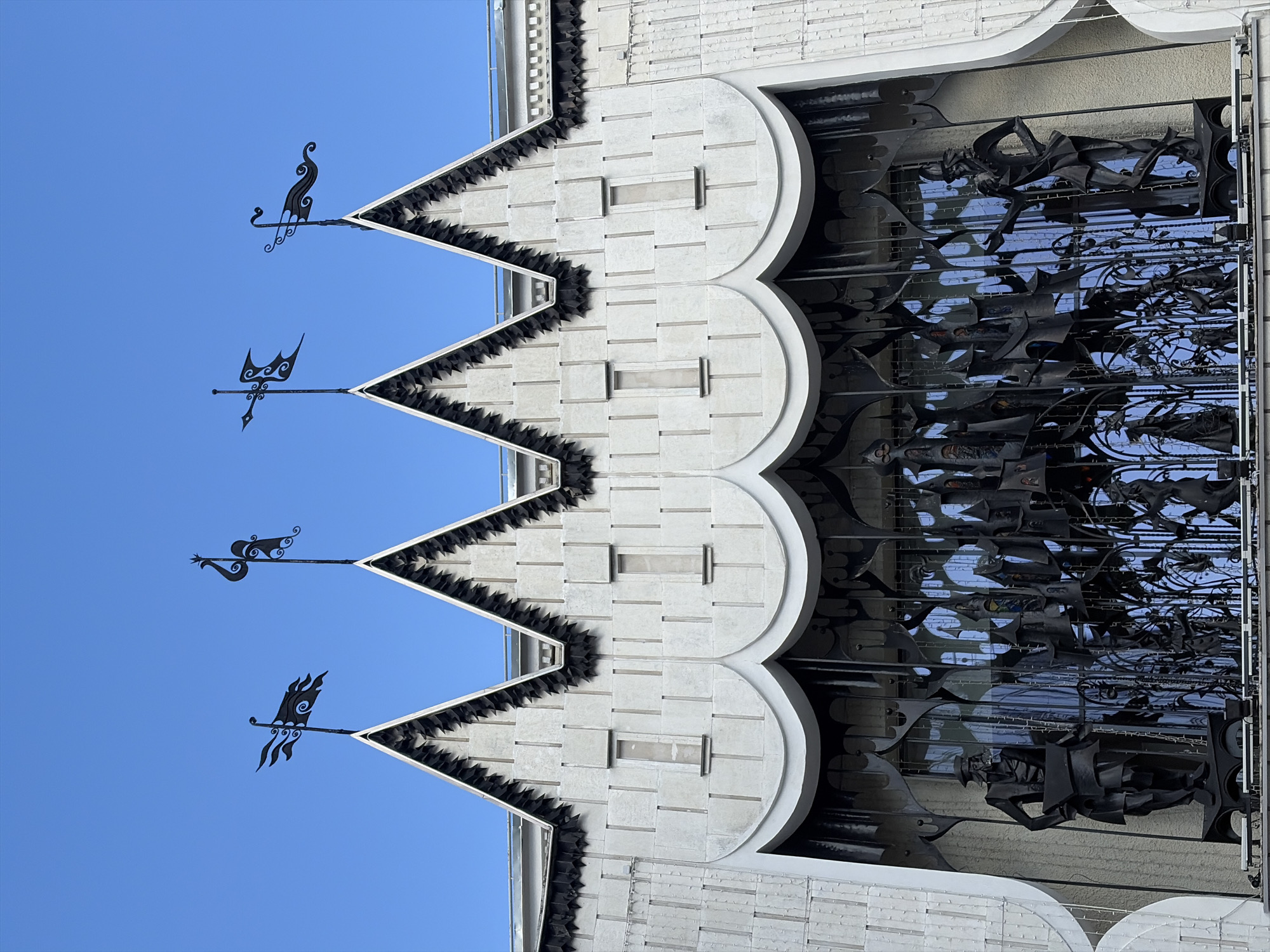 The image shows the facade of a building in Nizhny Novgorod, Russia. The building's upper section features a series of pointed gables, each topped with a decorative weather vane shaped like a flag or a bird. The facade is constructed from light-colored stone or concrete with vertical and horizontal grooved patterns. Below the gables, there are four narrow rectangular windows. The lower section of the facade is characterized by a series of curved, scalloped architectural elements. Dominating the foreground is an elaborate, black metal grate or sculpture. This metallic artwork depicts various figures, including what appears to be a man in a hat and costume on the left, holding a large piece of paper or fabric. Other figures, possibly in theatrical attire, are visible within the intricate metalwork, which also incorporates stylized architectural elements like towers and spires. The background visible through the grate is a bright blue, likely the sky. The overall architectural style suggests a historical or theatrical building.