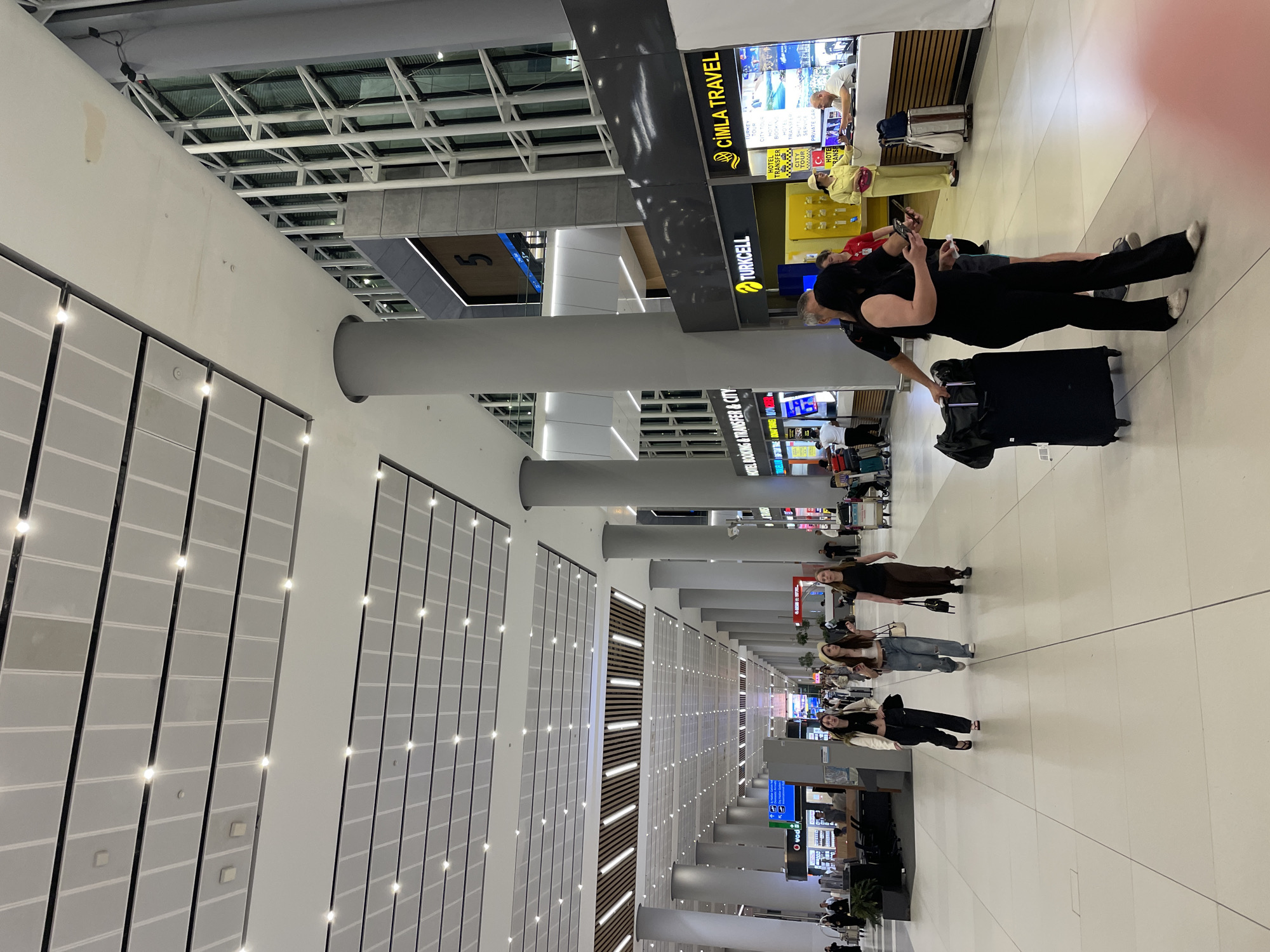 The image depicts a brightly lit, modern airport terminal interior. The ceiling is a complex structure of modular panels with integrated lighting, and large, cylindrical grey pillars support the elevated sections. The floor is made of large, light-colored tiles with visible grout lines.
In the foreground, a couple is walking towards the viewer. The woman, on the right, is wearing a black sleeveless jumpsuit and is holding her phone. The man, to her left, is partially obscured, wearing a dark shirt and black pants, and is also holding a phone. A large black suitcase with wheels is positioned behind the woman.
Further into the terminal, three young women are walking away from the camera. The woman on the left wears a white jacket over a black top and black pants. The middle woman has a light-colored hat, a white crop top, and light blue jeans. The woman on the right wears a black t-shirt and brown pants.
To the right of the central walkway, there are several storefronts. One prominently displays "TURKCELL" and another "ÇİMLA TRAVEL". The travel agency has signage advertising "HOTEL TRANSFER", "CITY TOUR", "HOTEL", "BUS", "TRANSFER", "SKI TOUR", and "PRIVATE CAR". Staff members are visible behind the counters of these establishments. In the background, more of the terminal stretches out, with numerous pillars and a distant ceiling extending into the distance. The overall impression is of a busy, functional transportation hub. The presence of "ÇİMLA TRAVEL" and the modern airport architecture suggest this is a commercial airport, possibly located in Arnavutköy, Turkey, given its size and amenities.
