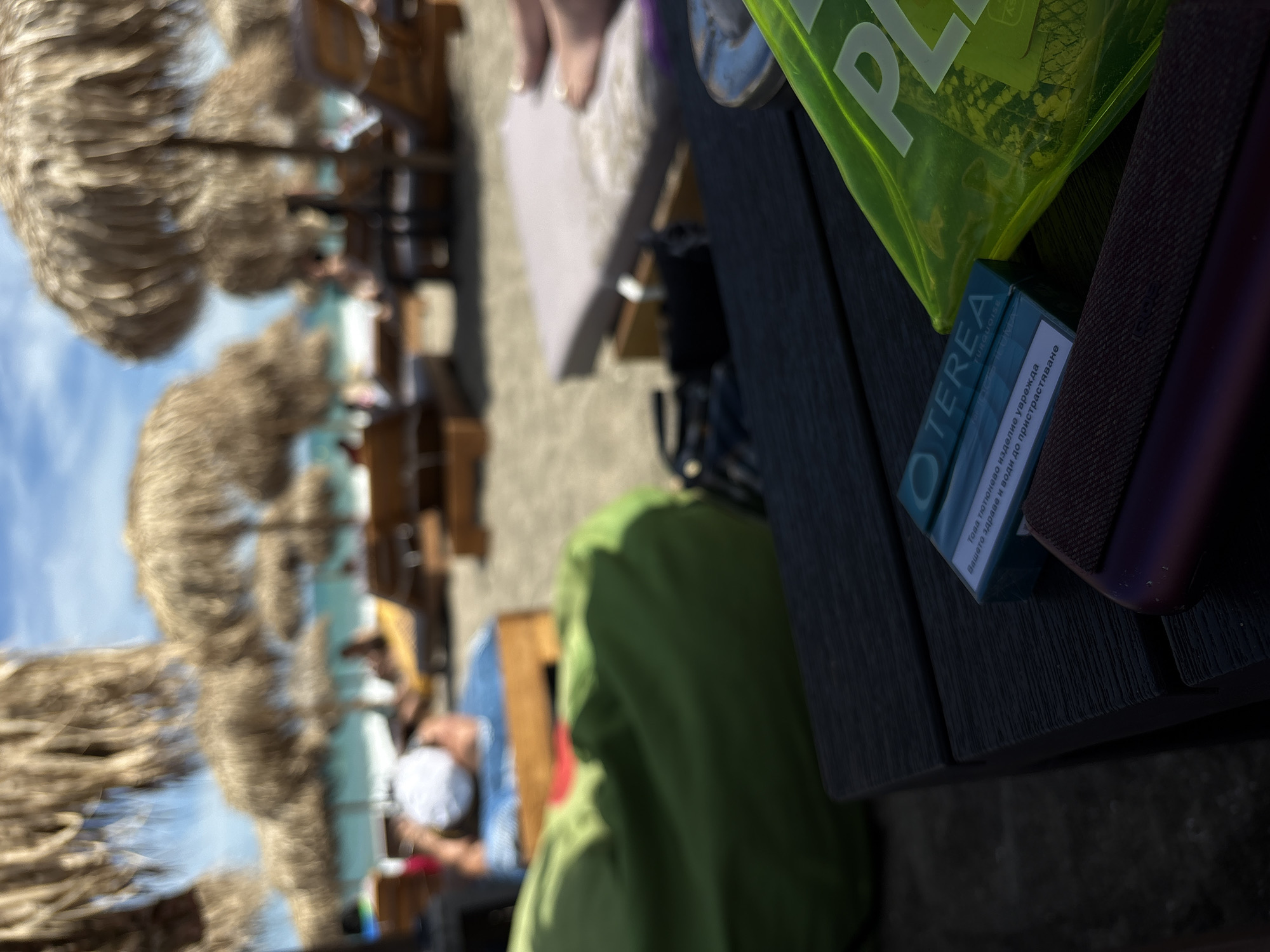 The image depicts a close-up view of a dark wooden table on a beach, with the blurry background showing a sunny day at the beach in Burgas, Bulgaria. There are several straw umbrellas and lounge chairs visible. In the foreground, on the table, are several items: a light blue and white package labeled "TEREA TURQUOISE" with some text in Bulgarian, a dark purple object, and a bright green translucent plastic bag with white lettering that appears to say "PL". A person's legs and feet, wearing white shoes, are visible resting on a lounge chair in the background, partially obscured by a large, crumpled green beanbag. The overall impression is of a relaxing day at the beach.