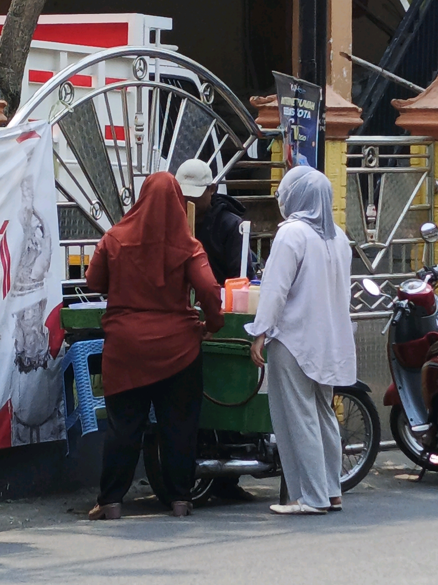 Two women stand near a food cart in Ketegan, Indonesia. The woman on the left wears a reddish-brown hijab and dark pants. The woman on the right wears a light gray hijab and light gray pants. Both women are facing away from the camera, towards the food cart, and appear to be interacting with it. The food cart is green and has a metal frame. Behind the cart, a white truck with red accents is partially visible. A motorcycle is parked to the right of the food cart. The background shows a building with yellow and brown colors and a metal fence. The scene appears to be taking place outdoors on a paved surface.