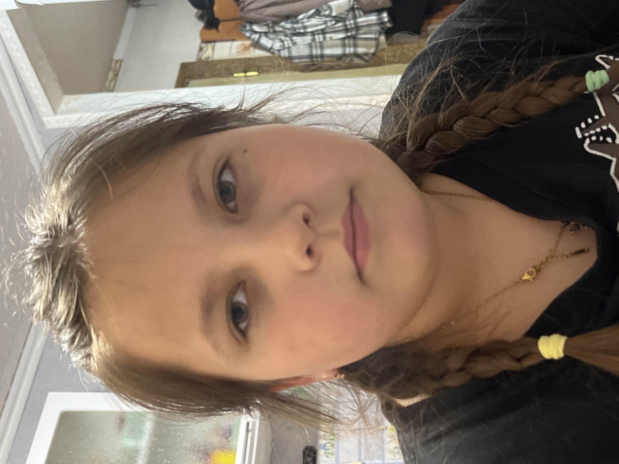 A close-up, slightly angled shot of a young girl with light brown hair styled in pigtail braids. The ends of the braids are secured with yellow elastic bands. She has fair skin, hazel eyes, and a neutral expression. She is wearing a black t-shirt and a gold necklace. The background is blurry and includes parts of a room with white cabinets and a glimpse of clothing on a shelf. The location provided, Novopokrovskaya, Russia, cannot be verified from the visual information.
