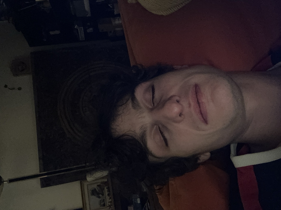 An image taken indoors in Vincennes, France, depicts a young adult male with fair skin and dark, wavy hair. His eyes are closed, and his mouth is slightly open, displaying a neutral to slightly pained or tired expression. He is positioned against a textured orange surface, likely a sofa cushion. A segment of his clothing, a dark garment with a white and red stripe on the collar/shoulder, is visible. The background is dimly lit and out of focus, revealing a floor lamp with a metallic pole, a framed picture on a surface to the left, and a dark shelving unit or furniture piece with indistinct objects to the right. A faint circular pattern is visible on a dark background element behind the subject's head.