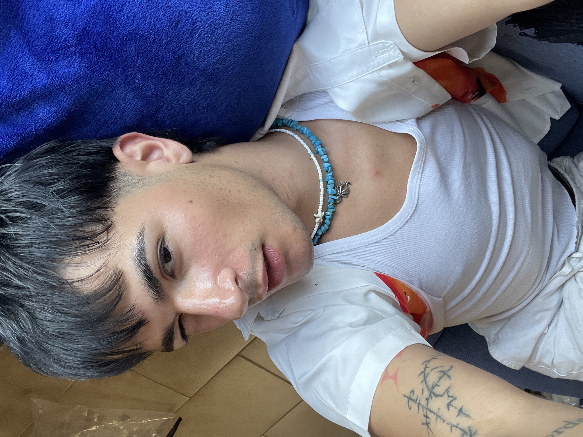 A young person with dark hair is depicted lying down in an indoor setting, likely in Su Masu/Elmas, Italy, bathed in bright natural light, suggesting it is daytime. They are looking towards the camera with a calm and relaxed expression.
The individual is wearing a white ribbed tank top layered under a white short-sleeved button-down shirt. The button-down shirt features an internal orange and red floral or abstract pattern, visible at the armpit and chest area. Around their neck, they wear a casual necklace composed of irregular blue turquoise-like beads interspersed with small white beads, featuring two silver marine-themed charms – one appearing to be a starfish and the other possibly an octopus or crab.
Their dark hair appears thick and slightly styled or damp. Subtle stubble is visible on their chin and jawline. A distinctive black ink tattoo with runic or abstract linear symbols is prominently displayed on their left forearm, accompanied by some red marks on the skin.
The person is resting their head on a dark blue plush pillow or fabric object, while their body lies on a light brown surface, possibly cardboard or a wooden floor. The overall scene conveys a sense of quiet relaxation. There is no visible text in the image.