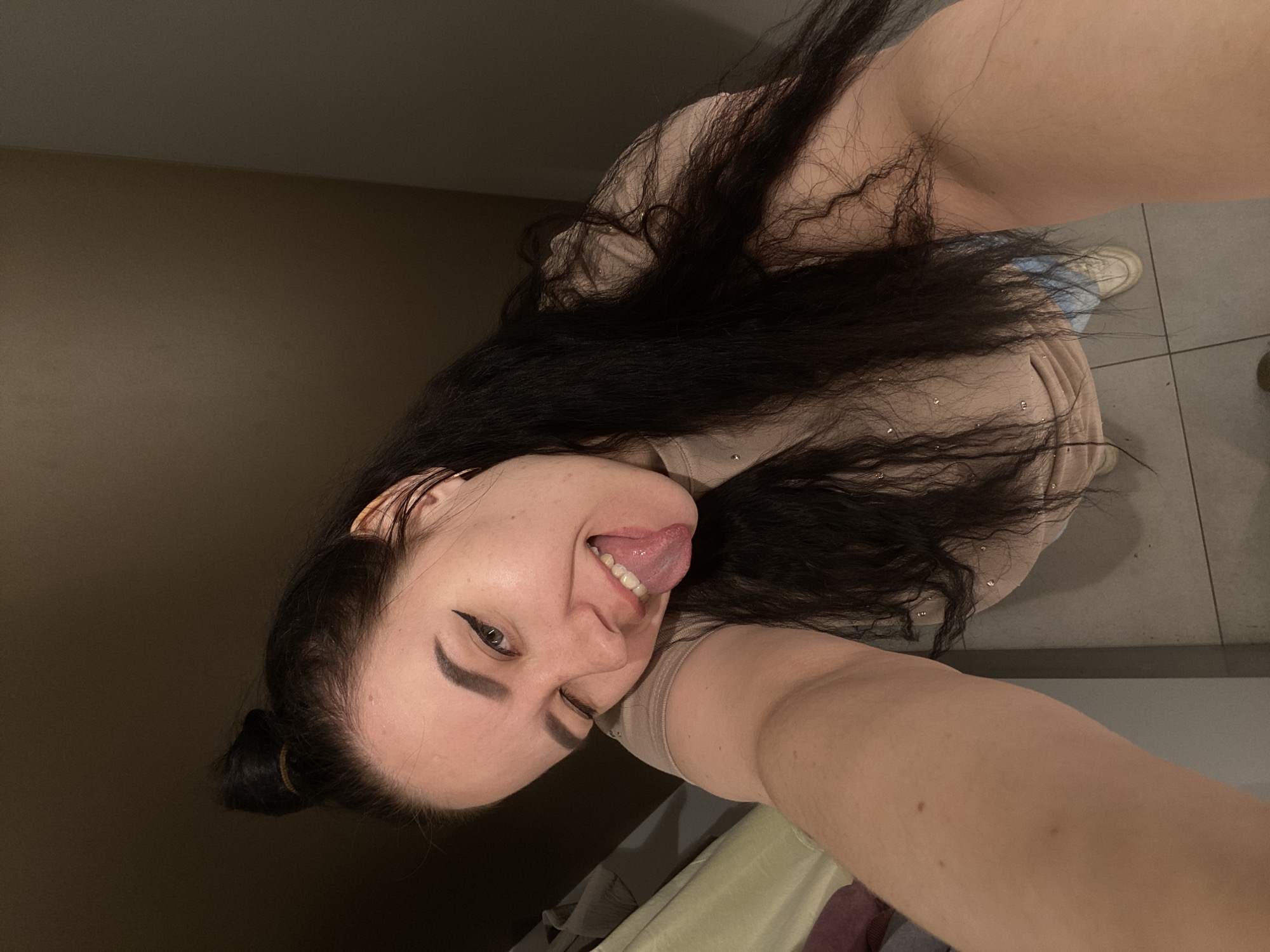 A woman with long, dark hair is taking a selfie indoors. She is sticking her tongue out and winking. She is wearing a beige shirt. Her hair is partially tied back. The background includes a brown wall and a tiled floor. The image may have been taken in a bathroom. It is not possible to discern whether the location is in Perm, Russia based on visual information alone.