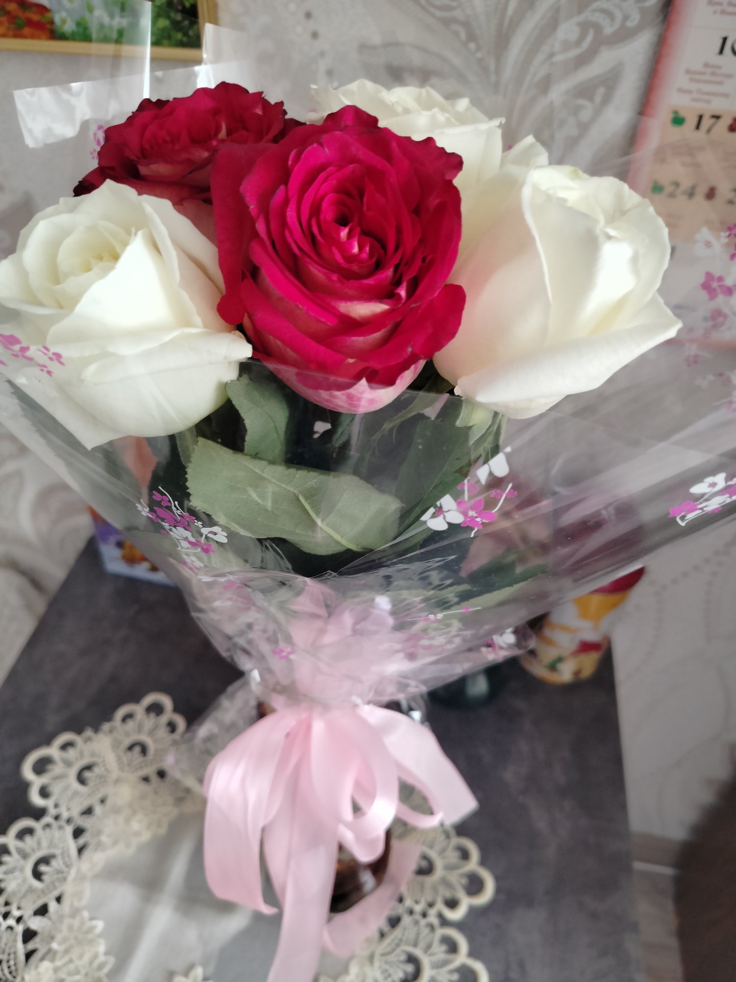 The image shows a bouquet of roses, with red and white flowers. The bouquet is wrapped in clear plastic with a pink floral pattern and tied with a pink ribbon. In the background, a calendar with Russian text is visible, along with a wall with a patterned wallpaper. A lace doily is placed on the left side of the image, and a dark surface, possibly a table, is visible below the bouquet. The scene appears to be an indoor setting, possibly a home or a shop, in Yashkino, Russia.