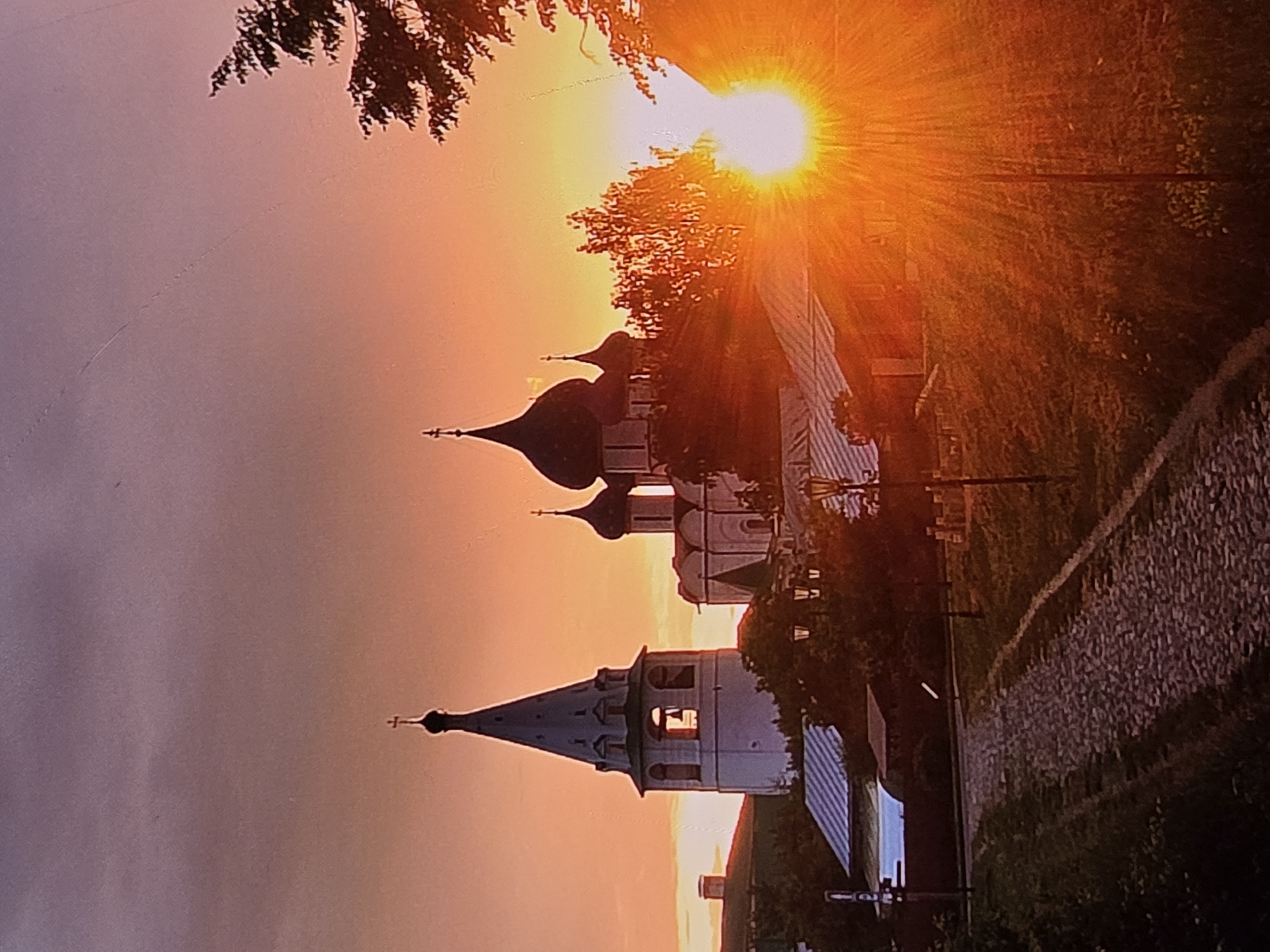 The image depicts a sunset in the city of Vladimir, Russia, with the Golden Gate and the Dormition Cathedral prominently featured. The sky is a vibrant gradient of orange, pink, and purple, with streaks of clouds. The Golden Gate, a historical monument, is visible on the left side of the frame, its white walls illuminated by the setting sun. The Dormition Cathedral, a magnificent church with onion domes, stands to the right, its dark silhouette contrasting with the bright sky. In the foreground, a cobblestone path winds its way up a hill, leading towards the historical structures. Trees and smaller buildings with red roofs are scattered throughout the scene, adding to the picturesque atmosphere. The sun, a brilliant orb, is setting behind the cathedral, casting a warm, golden light over the entire landscape and creating a dramatic effect with its rays.