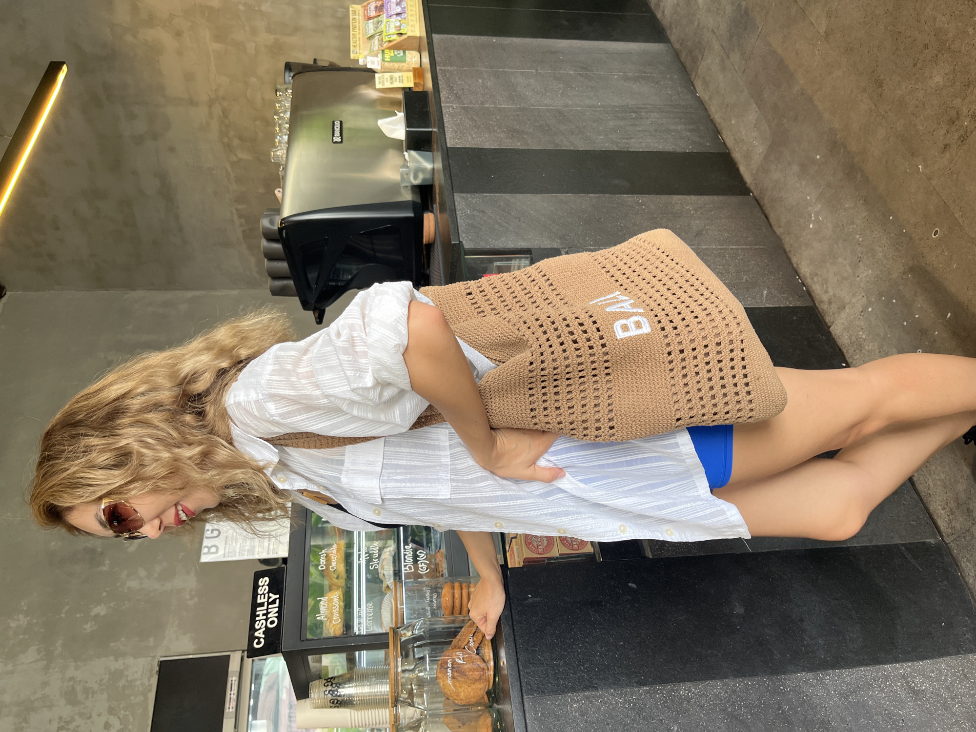 A woman stands in a cafe in North Kuta, Indonesia, facing left and looking away from the camera. She has blonde, wavy hair and is wearing oversized white sunglasses and a loose white button-up shirt over a blue garment. A brown crocheted tote bag with "BALI" written in white letters hangs from her shoulder. She is standing in front of a counter with baked goods displayed in glass jars and a coffee machine behind her. The cafe has a modern aesthetic with concrete walls and a black counter.