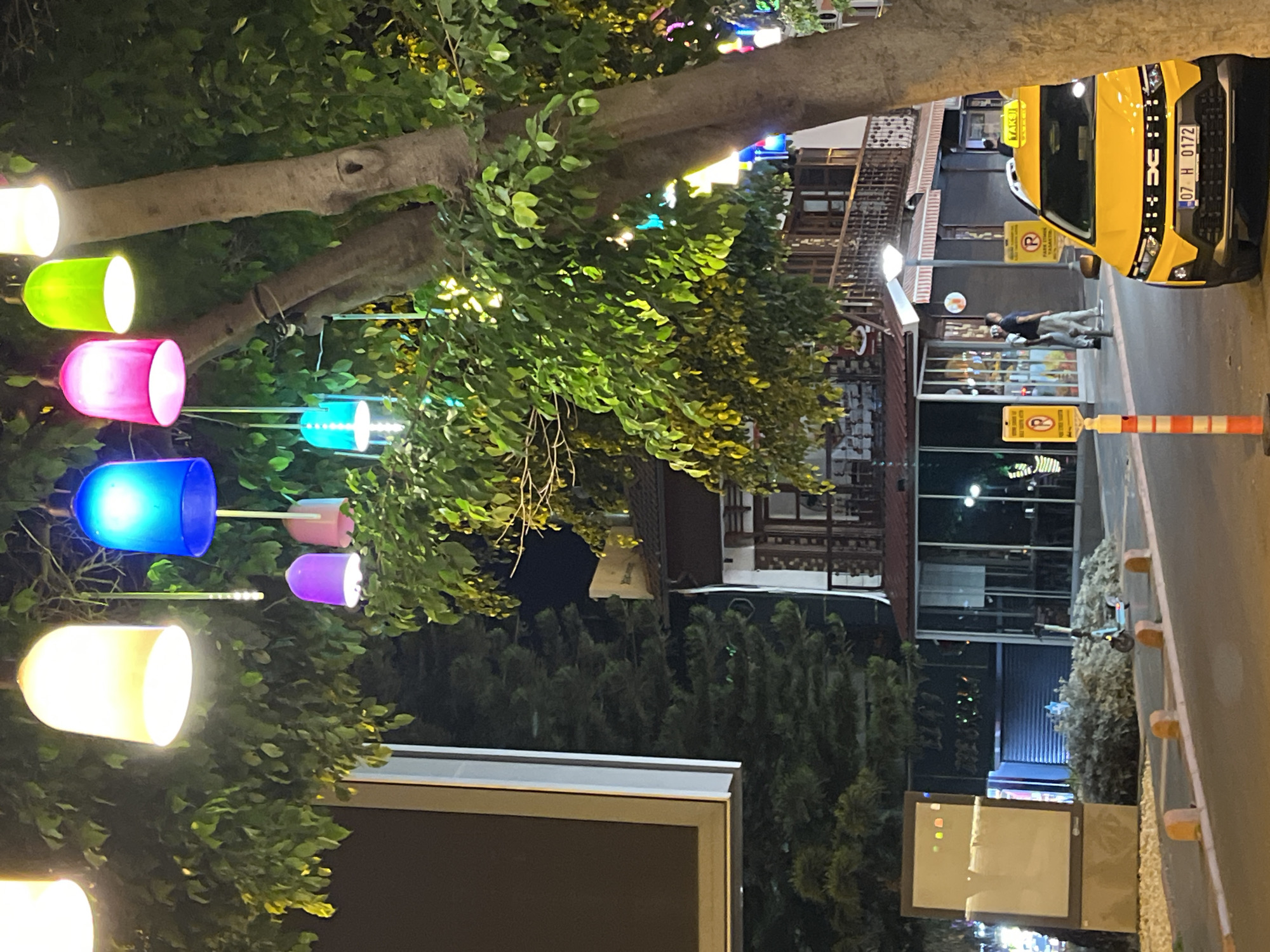 The image displays a vibrant night scene on a street, with the original image being rotated 90 degrees counter-clockwise. When viewed correctly, the foreground on the left features a large tree with lush green foliage, decorated with numerous glowing, cylindrical lights in various colors including white, bright green, pink, blue, purple, and yellow.
Further into the scene, a paved street or sidewalk is visible. A bright yellow taxi cab is prominent, either parked or slowly moving, with its front facing the viewer. The taxi's license plate "07 H 0172" is clearly visible, along with the "TAKSİ" sign on its roof. It features additional small lights on its front grille. A person wearing a dark shirt and light pants is walking on the sidewalk near the taxi.
Buildings with multiple stories, windows, and various lights line the street, suggesting shops or residences. One building has a red and white striped awning. Two yellow signs, each marked with a crossed-out "P" (likely indicating no parking) are visible. An orange and white striped bollard stands by the curb. An electric scooter is parked further down the street. The entire scene is illuminated by artificial lights, indicating it is nighttime. The overall atmosphere appears lively and urban, consistent with a commercial or tourist area in Alanya, Turkey.