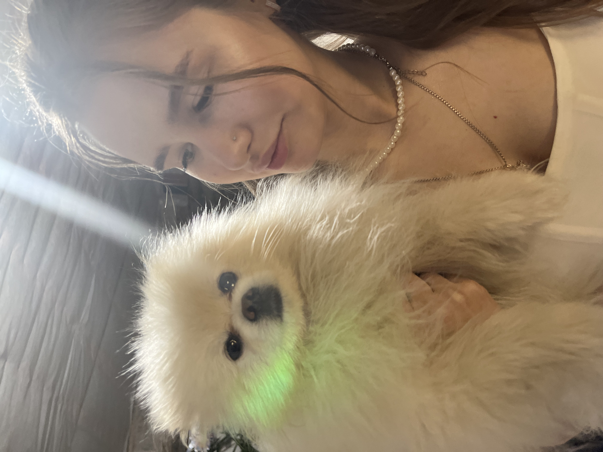 A woman is holding a white dog. The woman has brown hair and is wearing a pearl necklace and gold chain. The dog is small and fluffy. There is a bright light source visible on the left side of the image. The location is unknown, and there is no discernible evidence of Izhevsk, Russia.