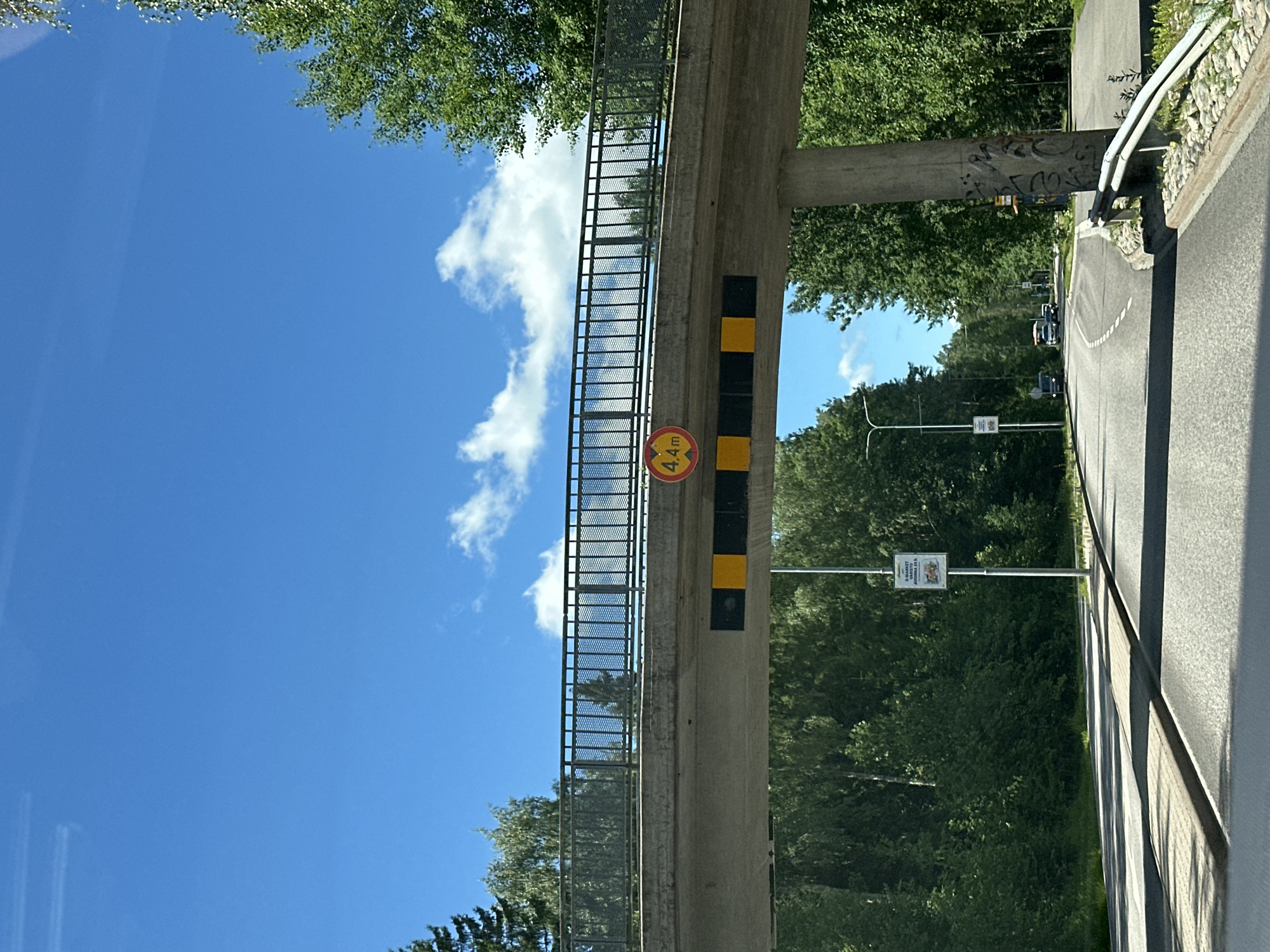 The image shows an overpass with a height restriction sign. The sign is circular with a red border and reads "4.4 m" in black text. Below the sign are alternating black and yellow stripes. The overpass is made of concrete and has a metal railing on the side. The scene is set outdoors on a sunny day with blue skies and some white clouds. Lush green trees line the road beneath the overpass. A few cars are visible on the road in the distance. The overall mood is calm and ordinary.