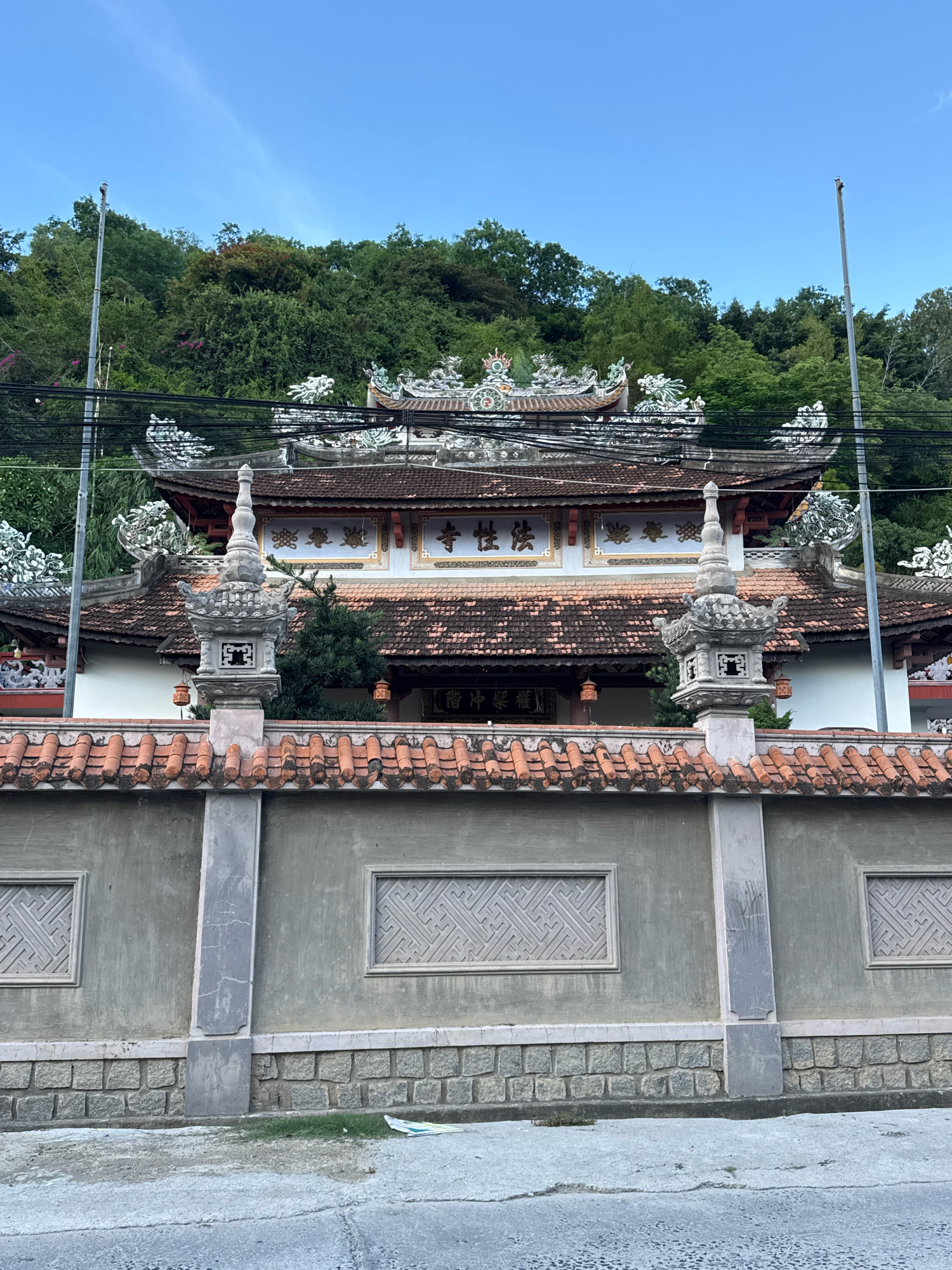 In Nha Trang, Vietnam, a multi-tiered building with a tiled roof, ornate architectural details, and multiple levels of eaves is visible behind a gray stone and concrete wall. The wall is topped with red tiles and features two rectangular panels with intricate geometric patterns. Two stone lanterns stand on pillars atop the wall. Electrical wires are strung across the upper portion of the scene. Behind the building, a lush green hillside covered in trees rises steeply, meeting a clear blue sky. The scene appears to be a temple or a similar cultural structure, set against a natural backdrop.