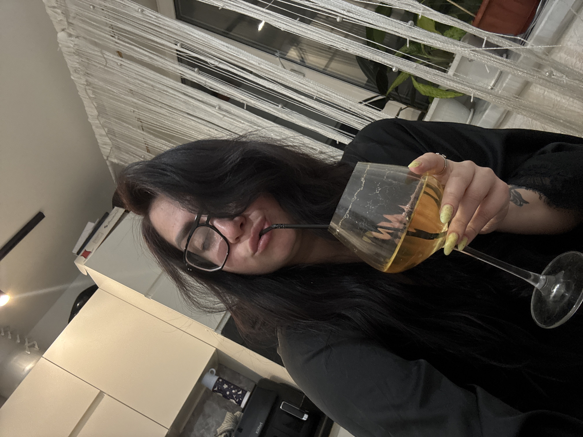 A medium, angled shot shows a woman with long, dark hair, wearing black-rimmed glasses, drinking from a wine glass with a straw. The wine glass contains a golden liquid, presumably wine, and is held by her hand with manicured fingernails. She is wearing a dark-colored garment.
In the background, a room is visible. A window is adorned with beaded strands, and a plant sits on the windowsill. A kitchen counter is also visible. The location is a city of Unknown, Unknown.