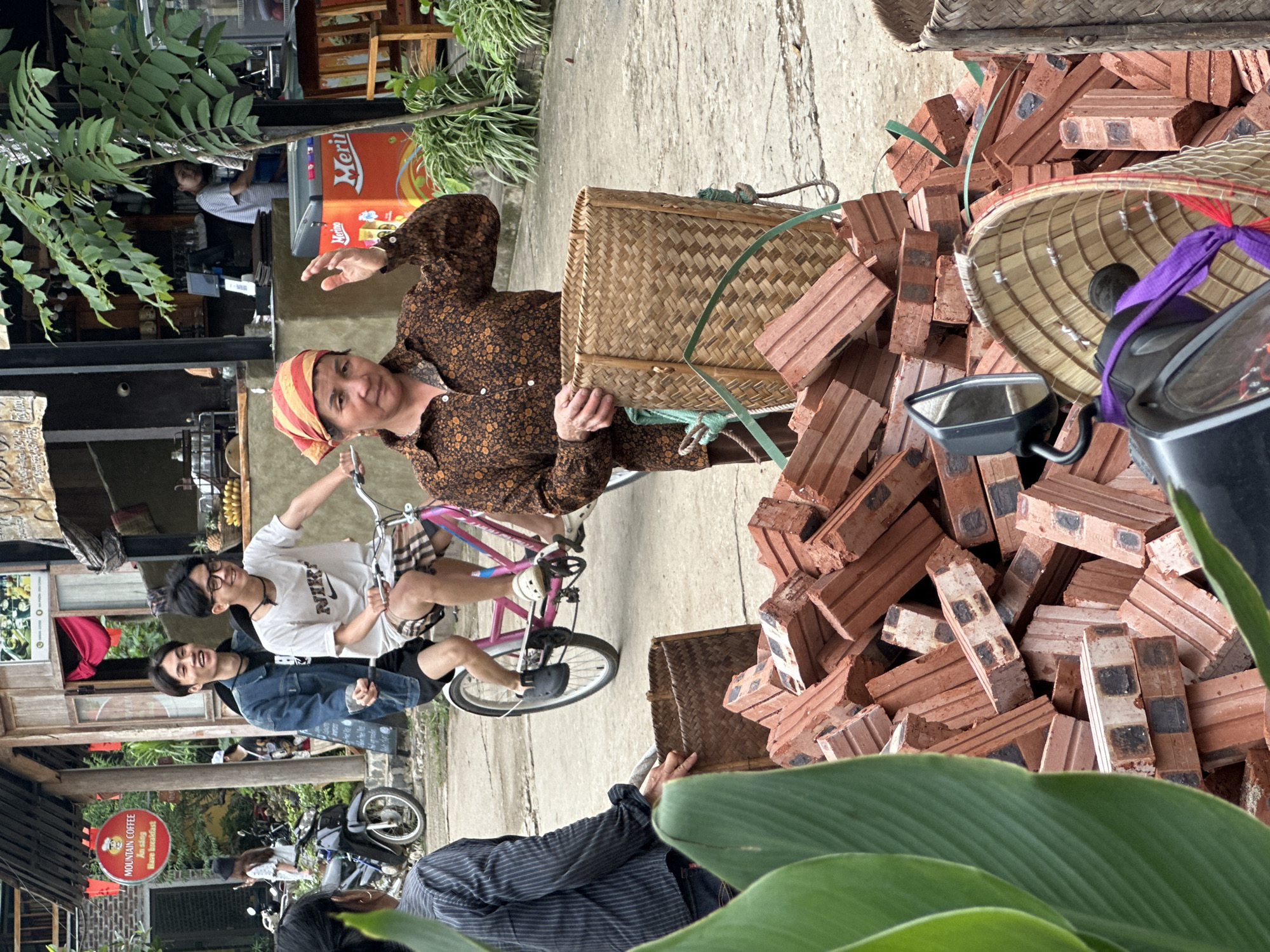 In Mai Chau, Vietnam, a woman wearing a brown patterned shirt and a red and yellow headscarf stands in the foreground, holding a woven basket and gesturing with her right hand. To her left, a pile of red bricks is visible, partially obscured by large green leaves. Behind her, two young men are riding a pink bicycle. The man in front, wearing a denim jacket, smiles, while the man behind him, wearing a white t-shirt and glasses, also smiles. In the background, buildings with wooden structures and signage are present. A motorcycle is parked to the left, and a bright orange cooler with the "Merino" logo is visible. Lush green plants and trees contribute to the outdoor environment. The scene appears to capture a moment of daily life in a Vietnamese town.