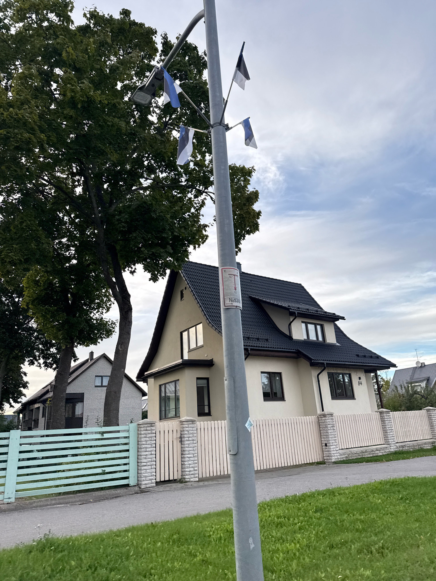 A street lamp post with multiple Estonian flags attached stands in the foreground, slightly off-center to the right. The flags are small and appear to be made of fabric. Behind the lamp post, a cream-colored house with a dark tiled roof is visible. The house has several windows with dark frames and a house number "84" is present on the exterior wall. A pale pink wooden fence with stone pillars separates the house from the street. To the left of the house, a large tree with green leaves is prominent. Further to the left, another house with a light-colored brick facade and a dark pitched roof is partially visible behind a light blue horizontal plank fence. The scene appears to be in a residential area of Tallinn, Estonia, with a paved road and a grassy area in the foreground. The sky is cloudy.