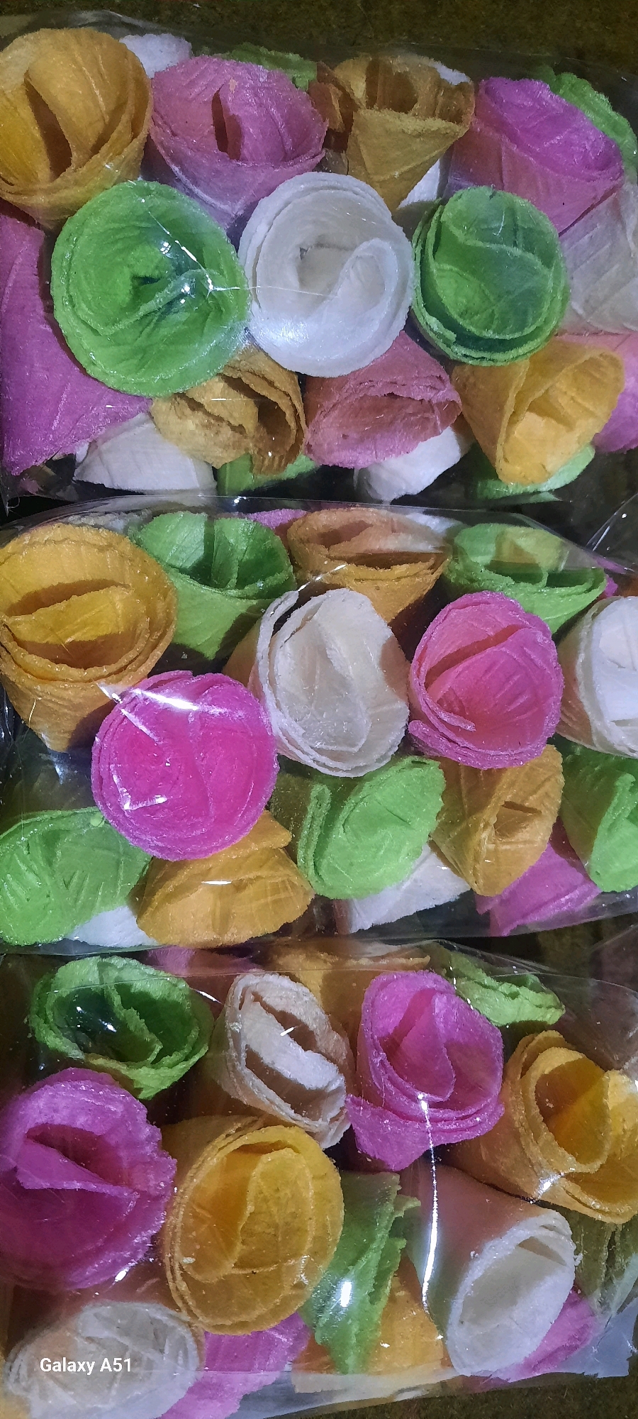 The image displays a close-up, overhead view of three clear plastic bags filled with an assortment of brightly colored, cone-shaped wafer snacks. The snacks are arranged in multiple layers, with individual pieces of yellow, pink, green, and white wafers visible. The wafers appear to have a textured, rolled surface, resembling flower petals. The lighting suggests an indoor setting, possibly a market or shop, given the presence of packaged goods. The background is softly blurred, but hints at a darker, possibly organic material. The bottom left corner of the image shows the text "Galaxy A51", indicating the device used for capture. There are no people or actions depicted in the image. The scene is entirely focused on the product itself. The location is identified as Ploso, Indonesia, suggesting these wafers are a local delicacy or product sold in that area.