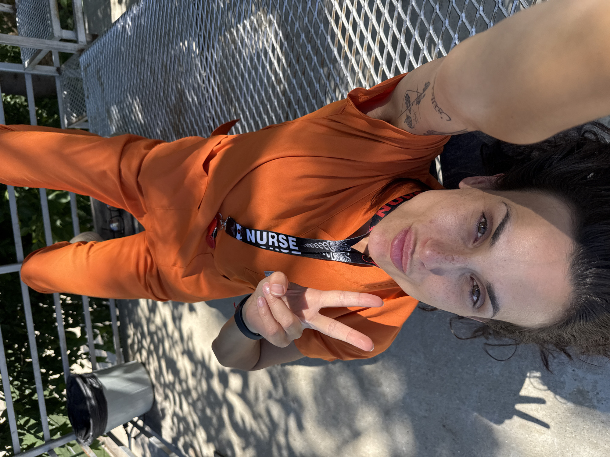 A person is lying on their back, holding a phone up to take a selfie. They are wearing orange scrubs and a black lanyard with the word "NURSE" printed on it. The person has dark hair and is looking at the camera with a slight smile. They are making a peace sign with their right hand.
The setting appears to be outdoors, with a metal grate in the background. There is also a trash can visible to the left of the person. The lighting suggests it is daytime, possibly late afternoon given the shadows. The overall mood seems relaxed and informal.
There is a tattoo on the person's left arm, which appears to be a design of arrows and other symbols. There are also some freckles visible on the person's face.