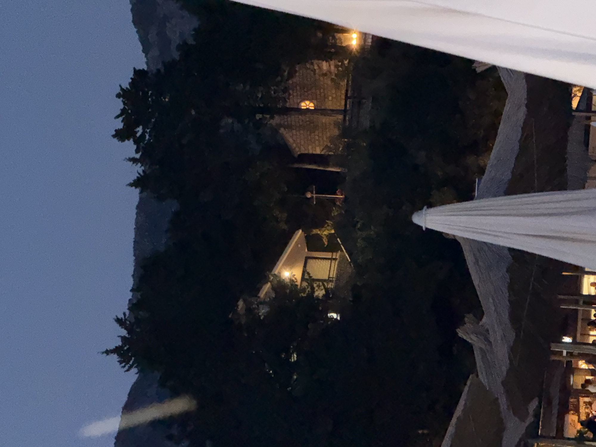 The image depicts a dimly lit outdoor scene, likely in Boreti, Montenegro, during twilight or early evening. In the foreground, several large white umbrellas and thatched-roof structures are visible, suggesting a beachside or outdoor seating area. Beyond these, a densely wooded hillside rises, with dark green foliage dominating the frame.
Nestled within the trees, two small buildings are discernible. The first, on the left, appears to be a structure with a light-colored facade and a pitched roof, illuminated by a warm light emanating from its windows. It seems to be elevated on a stone base. To its right, a stone building with a rough-hewn texture is visible, also featuring a lit window with a distinctive, possibly decorative, circular or star-shaped pattern. A thin, cross-like structure stands to the left of this stone building.
Further up the hillside, silhouetted against the darkening sky, are the shapes of trees and the contours of a mountain range. A bright, elongated light beam, possibly from a spotlight or a vehicle's headlights, streaks across the upper left portion of the image, illuminating a section of the distant hillside. The overall atmosphere is subdued and tranquil, with artificial lights creating small pockets of warmth against the encroaching darkness.